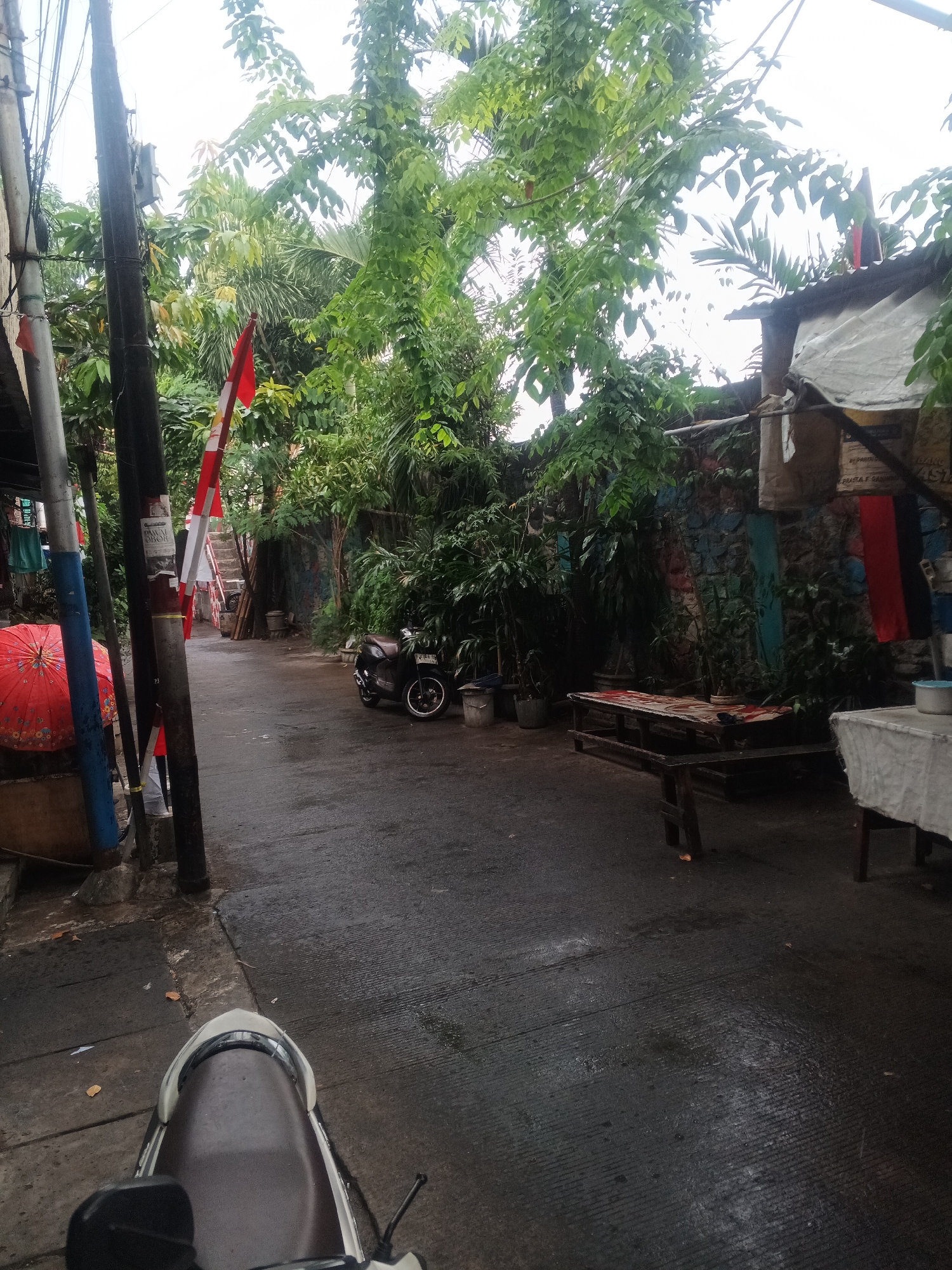 The image shows a narrow, wet street in West Jakarta, Indonesia, with dense greenery and several structures along the sides. A motorcycle is parked on the right side of the street, and a red umbrella is visible on the left. The scene appears to be an alleyway or a less-trafficked street in a residential area. The wetness of the road suggests it might have recently rained. Flags are displayed on a pole on the left side of the street. A wooden bench is situated on the right side of the street. The overall impression is one of a somewhat cluttered but lived-in urban environment with ample vegetation.
