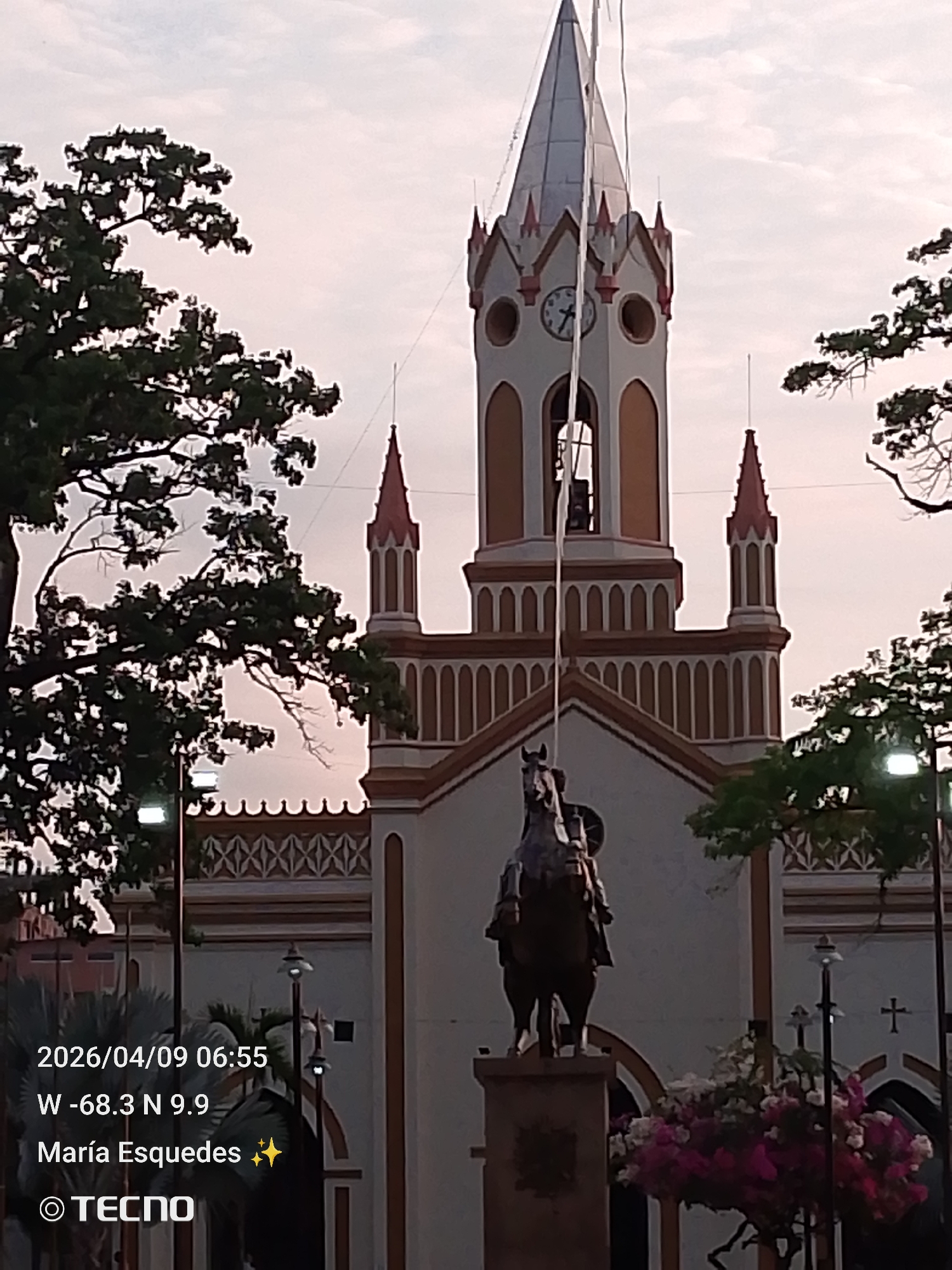 The image shows a building with a prominent tower and a clock. The tower is painted in white and orange, with red accents on its top. There are two spires on each side of the tower, in the same colors. A statue of a person on horseback stands in front of the building, and trees are framing the image on both sides. Streetlights are visible in the foreground, along with flowers. The image includes date, time, and GPS coordinates, indicating a location in María Esquedas, Tinaquillo, Venezuela.