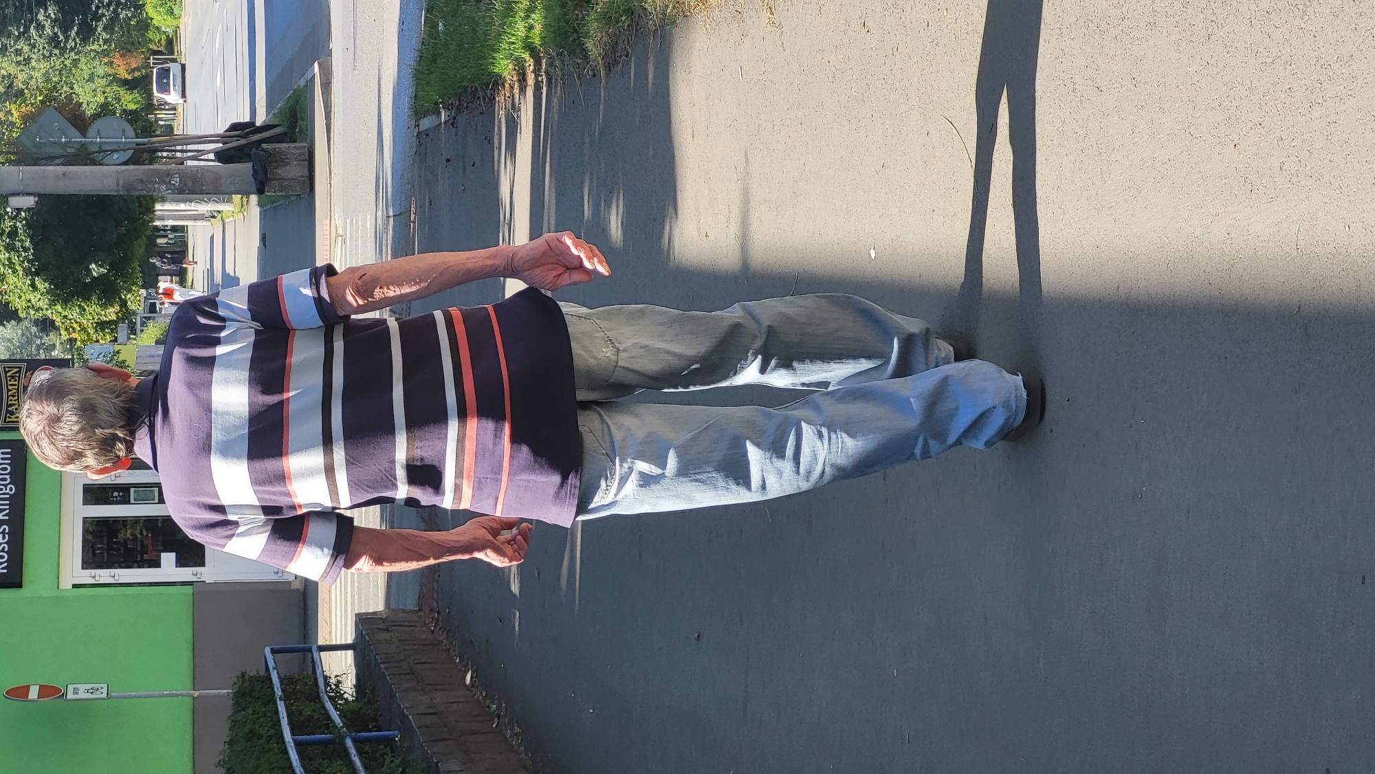 An elderly man with gray hair walks away from the camera on a paved sidewalk. He is wearing a horizontally striped short-sleeved shirt in navy blue, white, and coral, and light-wash blue jeans. His arms are partially visible and appear thin. The sidewalk is in a city setting, with a green building to his left that has "Roses Kingdom" and "KARMEN" signs visible. There is a street to the right of the sidewalk, with trees lining the street and a few cars in the distance. The sun casts a sharp shadow from the man onto the sidewalk. The location is identified as Prešov, Slovakia.