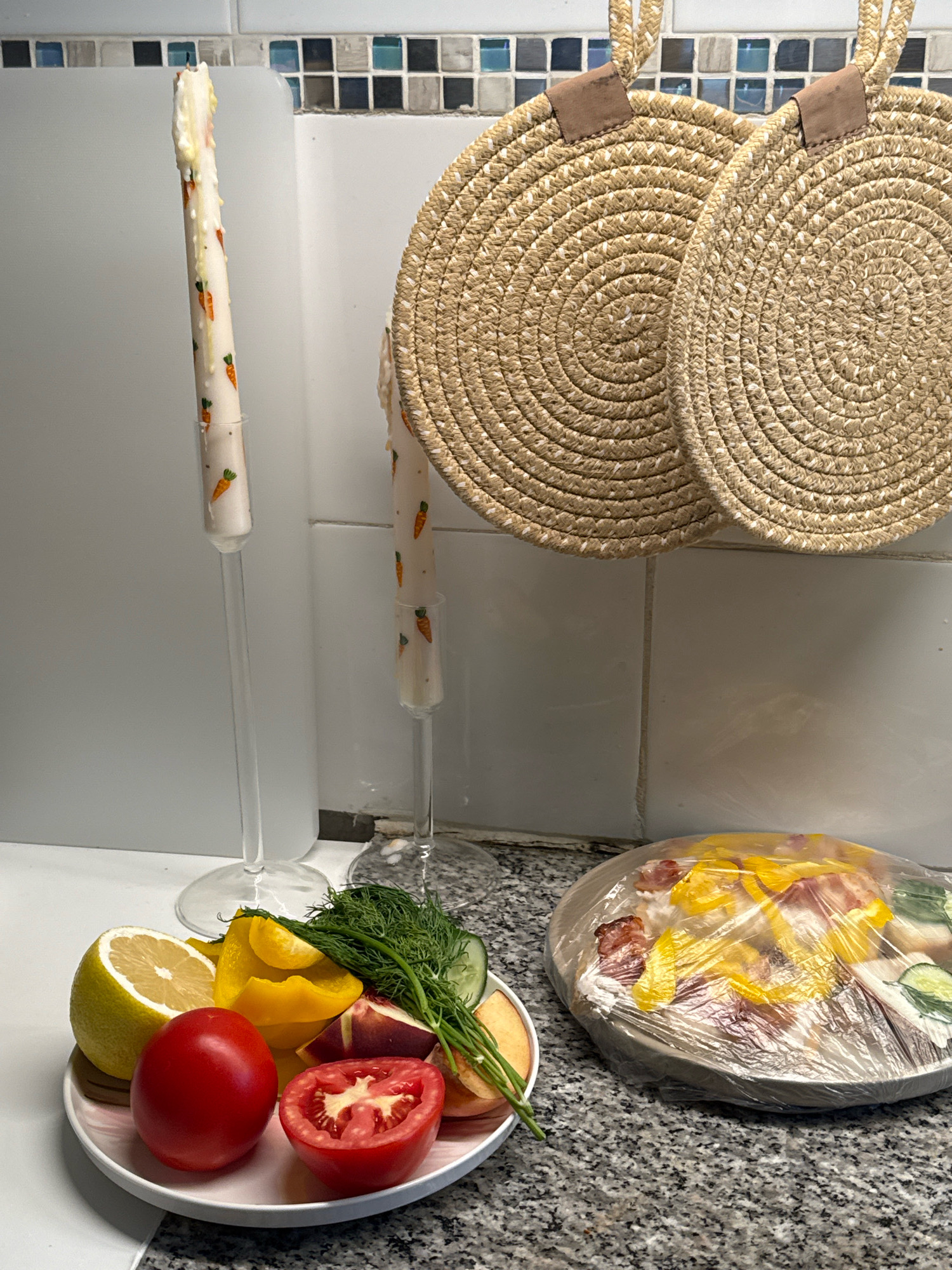 An eye-level shot captures a kitchen counter with various items, suggesting meal preparation or a decorative arrangement. In the foreground, a white plate holds an assortment of colorful ingredients: a bright red tomato, a halved tomato revealing its juicy interior, a bright yellow lemon wedge, chopped yellow bell pepper, a bunch of fresh dill, and slices of cucumber and peach. To the right of the plate, a round, flat dish is covered with transparent plastic wrap, obscuring its contents but hinting at a meal or ingredients within, possibly including bacon and yellow peppers. In the background, two tall, thin candles with a carrot pattern are displayed in clear glass holders, their wicks showing signs of being lit. Behind the candles, two round, woven placemats hang from the wall, their natural straw color contrasting with the white tiled backsplash. The scene is lit from above, casting soft shadows. The overall impression is one of a domestic setting, possibly a home kitchen, with elements that combine food preparation and decorative accents.