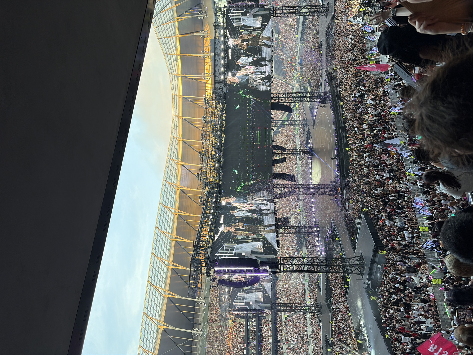 The image depicts a large-scale concert taking place in a stadium, likely in London, United Kingdom, given the architectural style of the stadium roof which resembles Tottenham Hotspur Stadium. The stadium is filled with thousands of spectators, many of whom are holding up lights or flags. The stage features multiple large LED screens displaying the performers, who appear to be a group of young men. The lighting is dramatic, with spotlights and stage effects illuminating the performers and parts of the crowd. Confetti is visible falling over the stage and parts of the audience, indicating a climactic moment of the performance. The overall atmosphere is one of excitement and a large, enthusiastic audience gathered for a major entertainment event.