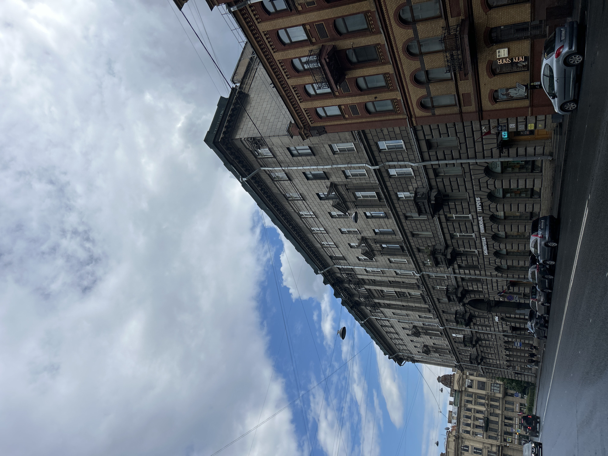 The image captures a street scene in a city, featuring a row of old, grand buildings lining a wet asphalt road. The sky above is partly cloudy, with patches of blue sky visible between the clouds. Overhead wires crisscross the sky, and a street lamp is suspended from one of them. The buildings are multi-story structures with detailed facades, typical of historical European architecture. The building on the left is a large, grey stone edifice with numerous windows and decorative elements. Adjacent to it is a building with a reddish-brown brick facade and arched windows, adorned with wrought-iron balconies. Several cars are parked along the street and a few people are walking on the sidewalk. The overall impression is of an urban environment with a rich architectural heritage.