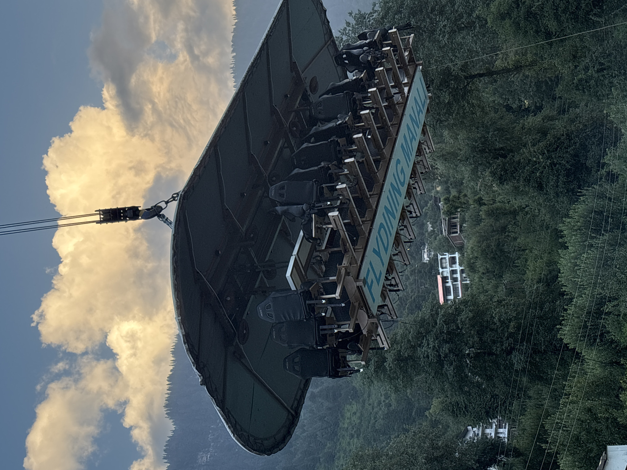 The image shows a "Dinner in the Sky" experience at Manali, India, suspended mid-air over a mountainous, forested landscape. A large, flat platform with multiple seats and tables is being hoisted by a crane. The platform has a dark, circular canopy overhead and is supported by a metal frame. The seats are black and appear to be bolted to the platform. A blue banner with the words "FLY DINING MANALI" is visible on the underside of the platform. Several people are seated at the tables, facing outwards. In the background, the lush green slopes of the Himalayas are visible, dotted with trees. A few buildings, possibly hotels or guesthouses, are perched on the hillsides. The sky is partly cloudy, with patches of sunlight illuminating the clouds. The overall impression is one of a unique dining experience set against a dramatic natural backdrop.
