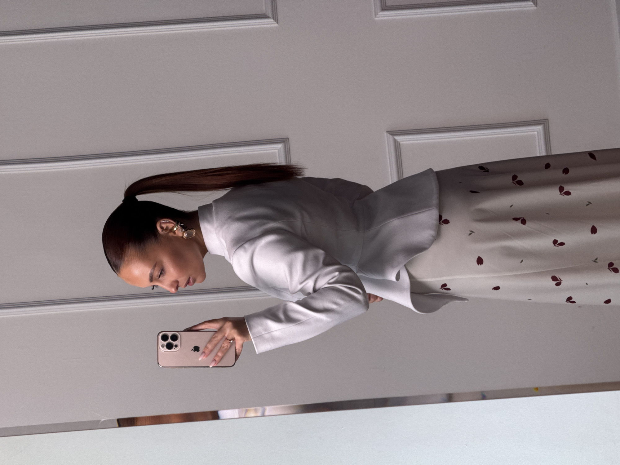 An individual is depicted taking a mirror selfie in an indoor setting within the city of Chaikovsky, Russia. The subject is an adult female, seen in profile facing right, with dark brown hair styled in a high ponytail. She wears a white, high-collared blazer or jacket with a structured, peplum hemline, paired with a light-colored skirt featuring a repeating pattern of small, dark red, abstract leaf or petal shapes. Her left hand is raised, holding a rose gold or champagne-colored smartphone, identified as an iPhone with a visible Apple logo and triple camera array. A ring is present on the fourth finger of her left hand, and her fingernails are manicured with light pink polish. Multiple golden-toned earrings are visible in her right ear, including a prominent rectangular-shaped one. The background consists of a white wall adorned with classical-style rectangular decorative molding panels. A portion of a mirror frame is visible on the far left of the image. The individual's gaze is directed towards the phone's screen or her reflection within the mirror.