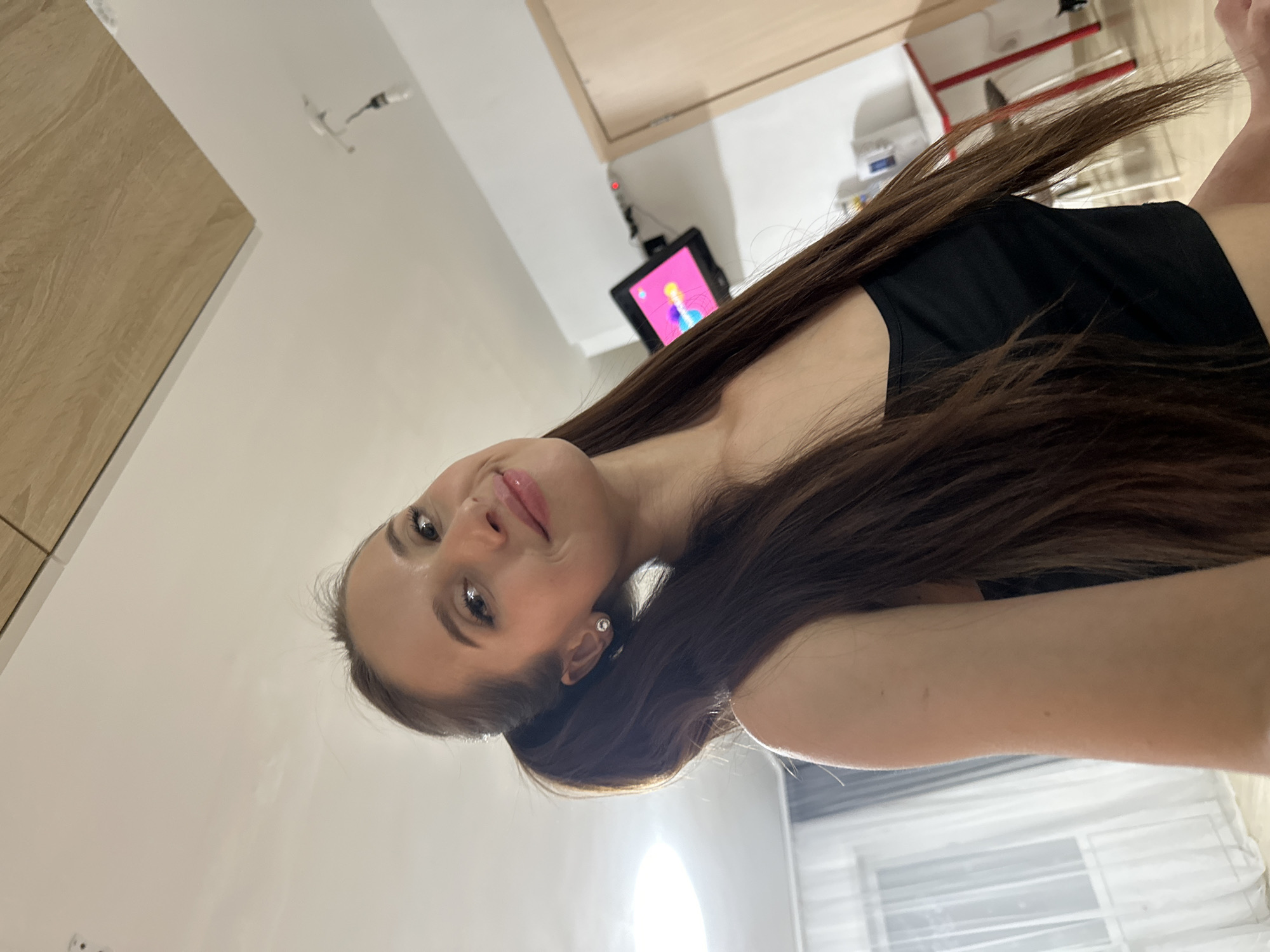 A woman with long brown hair is depicted in an indoor setting. The camera angle is tilted, offering a close-up view of her face and upper body. She is wearing a black top. A screen with a pink background is visible in the background. The background also includes furniture and a window. This image does not provide specific details to identify the location as Yekaterinburg, Russia.