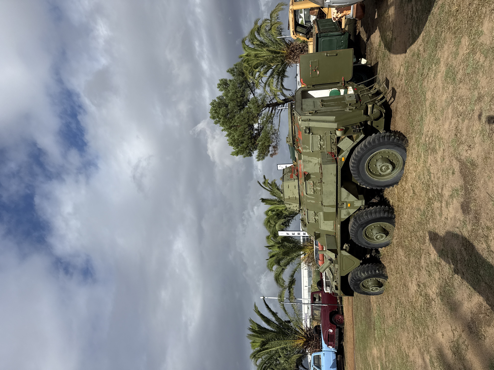 The image shows an outdoor scene in Kraaifontein, South Africa. Dominating the view is a line of vintage military vehicles parked on a grassy field. In the foreground, the prominent vehicle is a large, olive-green armored personnel carrier with six wheels. Behind it are several older civilian vehicles, including a red pickup truck and a blue truck. Scattered palm trees are interspersed amongst the vehicles and along the background. The sky above is overcast with dark storm clouds.