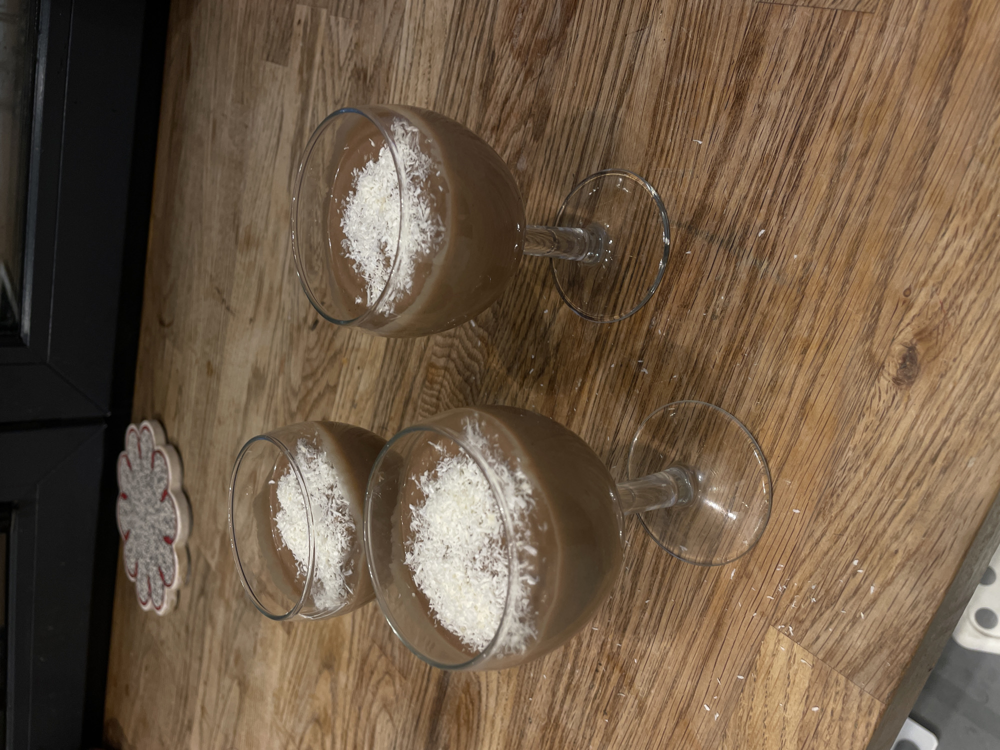 Two dessert glasses containing a brown liquid topped with shredded coconut are on a wooden surface. The glasses have a stemmed base and a clear bowl shape. Beside the glasses is a decorative trivet. The location is not identifiable as Moscow, Russia.