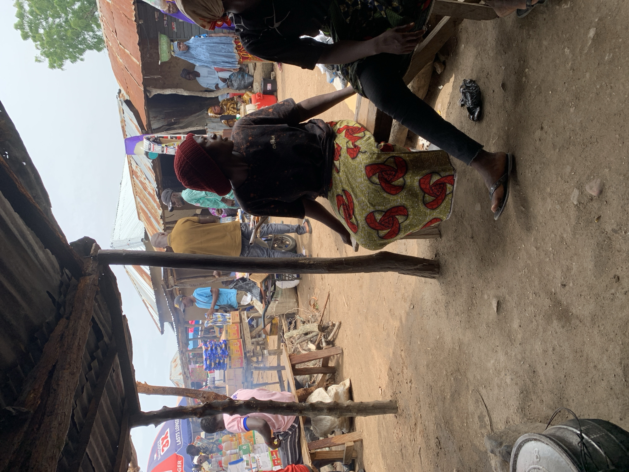 The image depicts an outdoor market scene in Dull A, Nigeria. In the foreground, two individuals are seated on a wooden bench. The person on the left wears a dark, graphic t-shirt and a vibrantly patterned skirt with red and green motifs. They are looking to the right. The person on the right is partially visible, wearing dark trousers and a patterned garment. Their legs are crossed, and they are wearing flip-flops.
Behind them, a bustling marketplace unfolds. Several stalls with corrugated tin roofs are visible, some appearing weathered. A variety of goods are displayed, including packaged items in blue and yellow boxes, and bottles on shelves. People dressed in traditional clothing are milling about. One person in a light blue garment is seen in the background, and another in a light green garment with a cap. There are also signs visible, including one with red and blue branding. The ground is predominantly dirt, with some scattered debris and wooden structures. The overall atmosphere suggests a lively, everyday market environment.