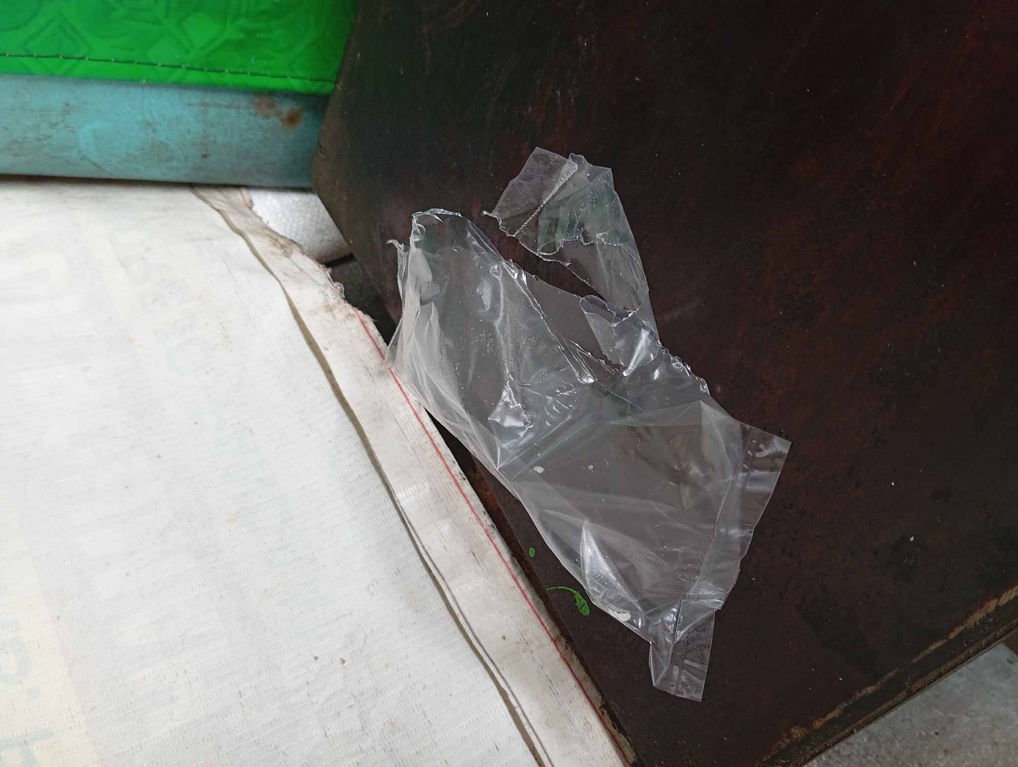 A crumpled, transparent plastic bag is on a dark wooden table. The bag is torn and appears to have been used. In the background, there is a white textured surface, a teal cylindrical object, and a green textured object. A thin red line is visible along the edge of the white textured surface. The image is taken from a slightly elevated angle, looking down at the table. The location is stated to be Lemahbang, Indonesia.