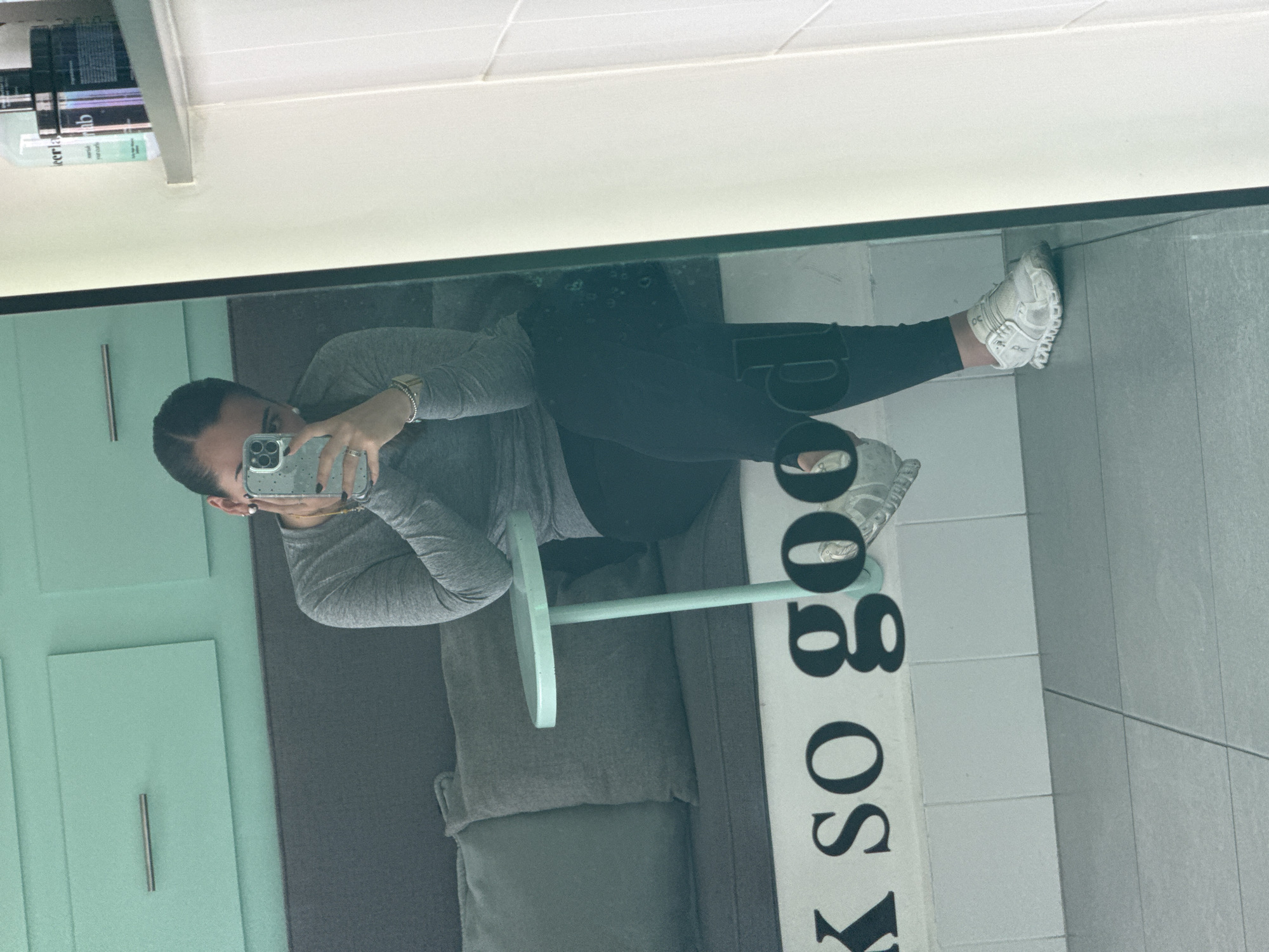 A person is captured in a mirror selfie, seen from the waist up, standing in what appears to be an indoor setting. The individual, a female, is dressed in a grey long-sleeved top and black leggings, with white athletic shoes. She wears a silver-toned watch on her left wrist and rings on her fingers. Her hair is pulled back, and she holds a smartphone with a patterned case up to her face to take the picture, reflecting her image in the large mirror that forms the foreground of the scene.
The background visible in the mirror reflection includes a light teal or mint green wall paneling with a silver handle to the person's left, suggesting a door or cabinet. To her right, a light mint green round table with a slender pedestal stands next to a grey upholstered bench or seating area. The floor is tiled with light grey and white sections. Above, a white ceiling with linear lighting is reflected, along with some shelves holding bottles, some with illegible text.
Prominently featured on the white section of the tiled wall, reflected in the mirror, are large, dark, reversed letters that read "OO SO X". When corrected for the mirror's reflection, this likely spells out "SO GOOD" or a similar phrase. The scene suggests a modern, well-lit indoor space, possibly a lounge, dressing room, or retail environment. The image is stated to be from San Bernardino Tlaxcalancingo, Mexico.