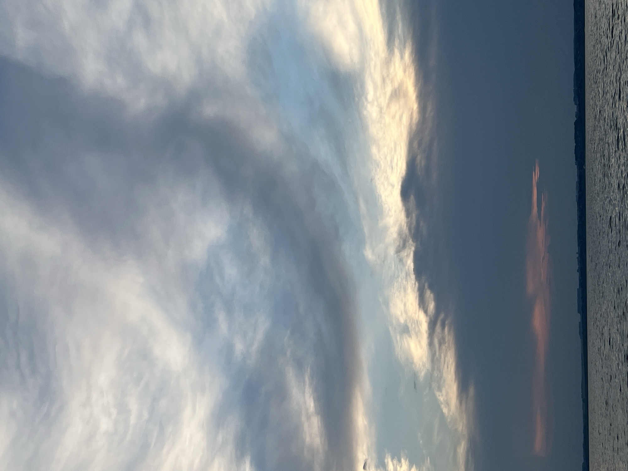 The image depicts a vast expanse of sky filled with a variety of clouds, ranging from wispy cirrus formations to thicker, darker stratus clouds. The sky transitions from a pale blue near the horizon to a more intense blue higher up, with patches of white and grey clouds scattered throughout. The lower portion of the image is dominated by a dark, ominous band of clouds that obscures the horizon. Below this band, a sliver of pinkish light suggests the presence of a sunset or sunrise. The bottom of the image shows a body of water, likely a lake or river, with a calm surface reflecting the muted colors of the sky. The distant horizon is lined with a dark, silhouetted landmass, possibly a treeline or low-lying buildings. The overall impression is one of a dramatic and moody sky over a serene body of water. The location is identified as Novopichugovo, Russia.