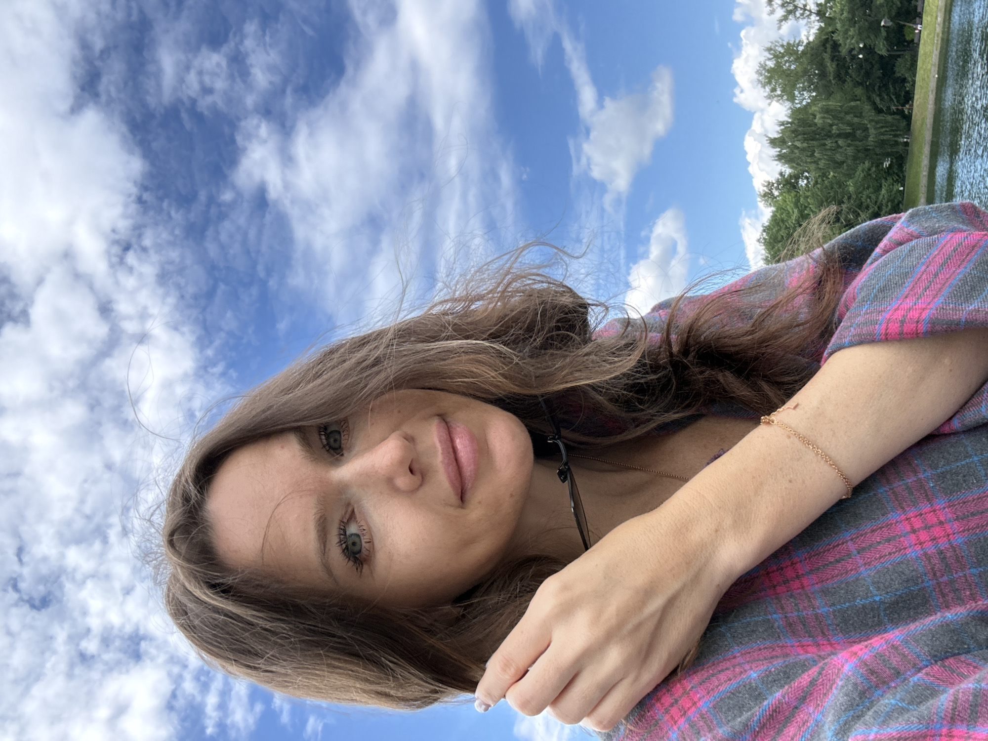 The image is a selfie taken outdoors, likely on a sunny day, with a bright blue sky and scattered white clouds in the background. The subject is a woman with long, brown hair, fair skin, and green eyes. She is wearing a pink and gray plaid shirt and a delicate gold bracelet on her left wrist. Her left hand is raised to her face, touching her hair, and she is holding a pair of dark-rimmed glasses. The background includes a body of water, likely a pond or lake, and a line of lush green trees. The overall setting suggests a park or recreational area, possibly in or near a city. The reflection in the water is not clear enough to discern specific details. The style of the trees and the overall ambiance could be consistent with various locations within Moscow, Russia, known for its numerous parks and waterfront areas.