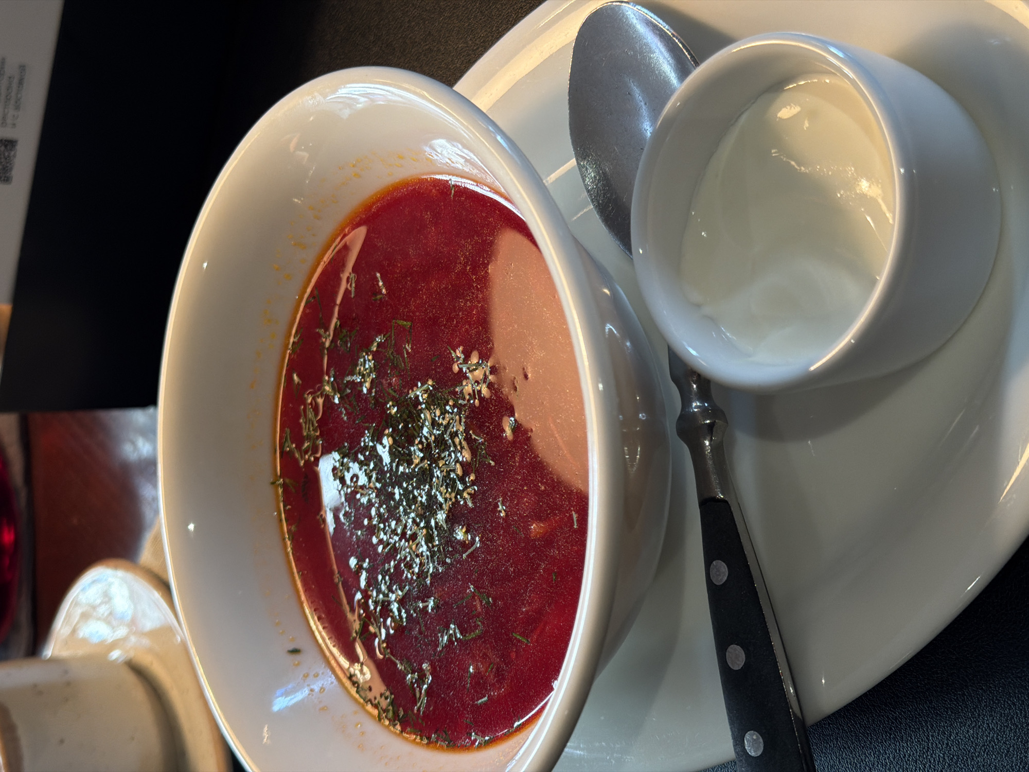 A close-up shot depicts a bowl of borscht garnished with dill, accompanied by a side dish of sour cream, and a spoon. The meal sits on a white plate. This image was taken at a restaurant in Krasnoyarsk, Russia.