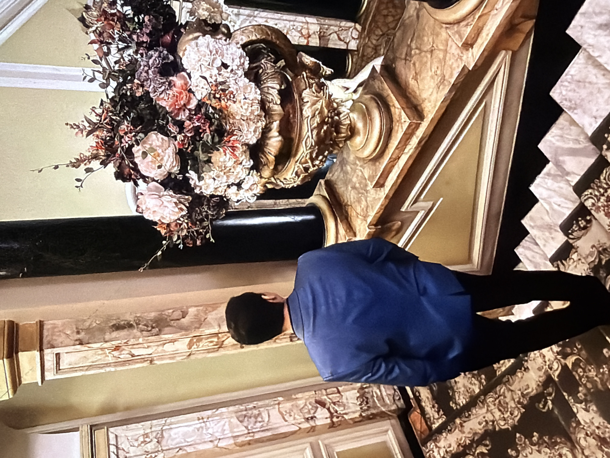 The image captures a high-angle view of a grand staircase inside the National Opera of Ukraine in Kyiv, Ukraine. A man, seen from behind, descends the stairs, wearing a dark blue blazer and black trousers. The upper visible section of the staircase is covered in a dark carpet with a light, intricate pattern, transitioning to lighter-toned marble steps further down. To the right, on a marble landing, a large, ornate golden urn sits on a matching marble pedestal. The urn is densely filled with an arrangement of artificial flowers in shades of pink, cream, red, and deep purple. Pale green walls frame the background, interspersed with vertical architectural elements featuring ornate, veined marble paneling in tones of pink, brown, and white, capped with golden accents. Dark, slender pillars are partially visible behind the floral arrangement. The lighting is bright, highlighting the opulent details of the interior.