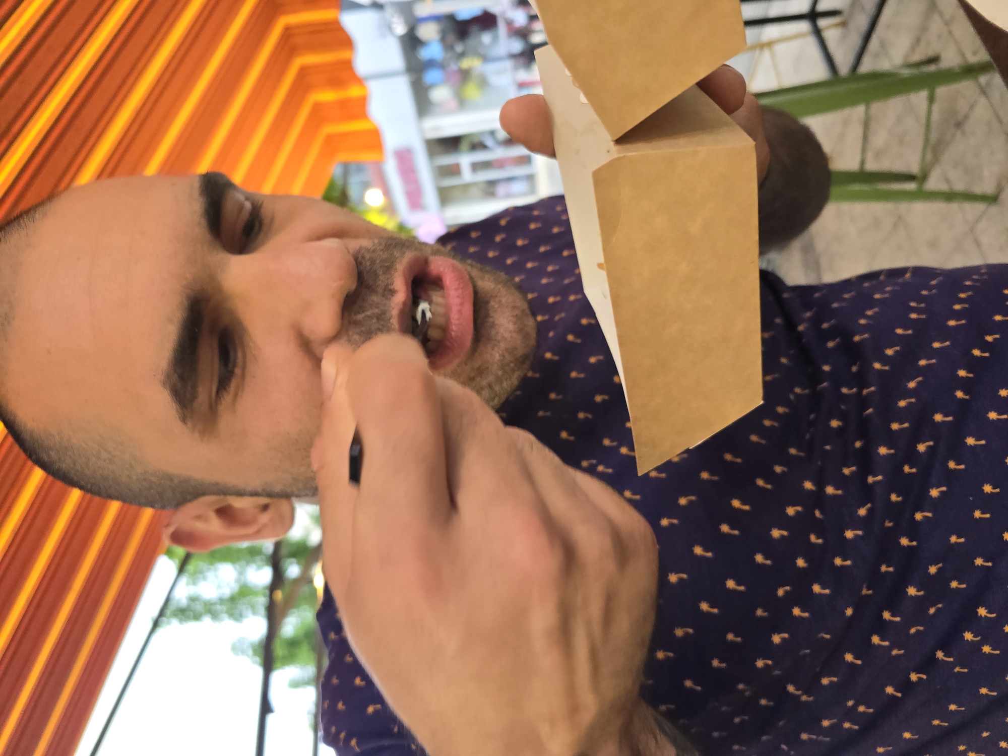 The image shows a man with a shaved head, wearing a patterned shirt, appearing to be picking something from his teeth with a small black object. He is holding a small brown box. The background appears to be a slightly blurry outdoor scene. Due to the lack of visual indicators, the location in Pleven, Bulgaria cannot be confirmed.