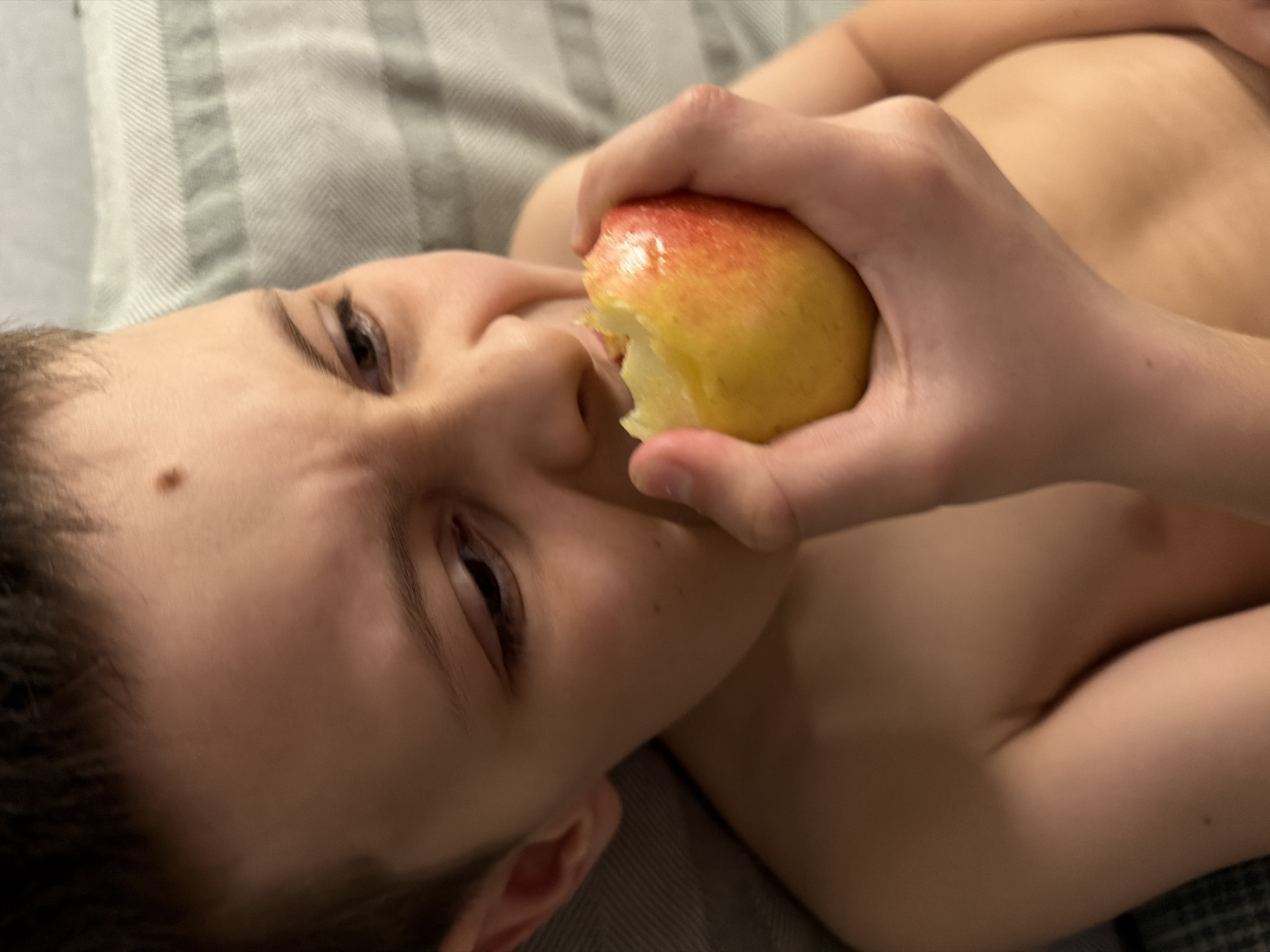 A young shirtless child is lying on a gray striped pillow, eating a yellow and red apple. The child is holding the apple in their right hand and taking a bite. The child has short, dark hair and is looking towards the left. There is a mole on their left cheek. It is impossible to determine the location of the image as Votkinsk, Russia with the available information.