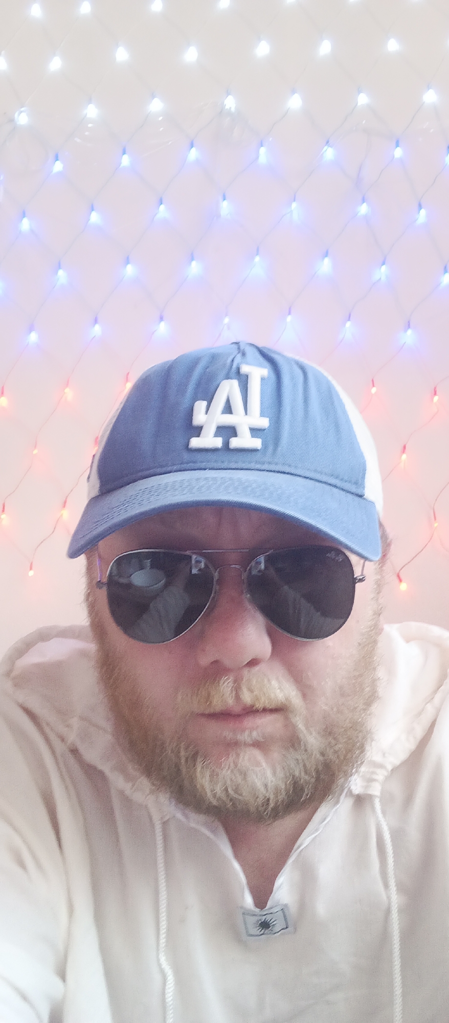 A man wearing a light blue and white Los Angeles Dodgers baseball cap and aviator sunglasses is shown in a close-up shot. He has a red beard and mustache and is wearing a light-colored hooded sweatshirt. The background consists of white and blue Christmas lights strung in a grid-like pattern, with some red lights interspersed. The location is Unknown, Unknown.