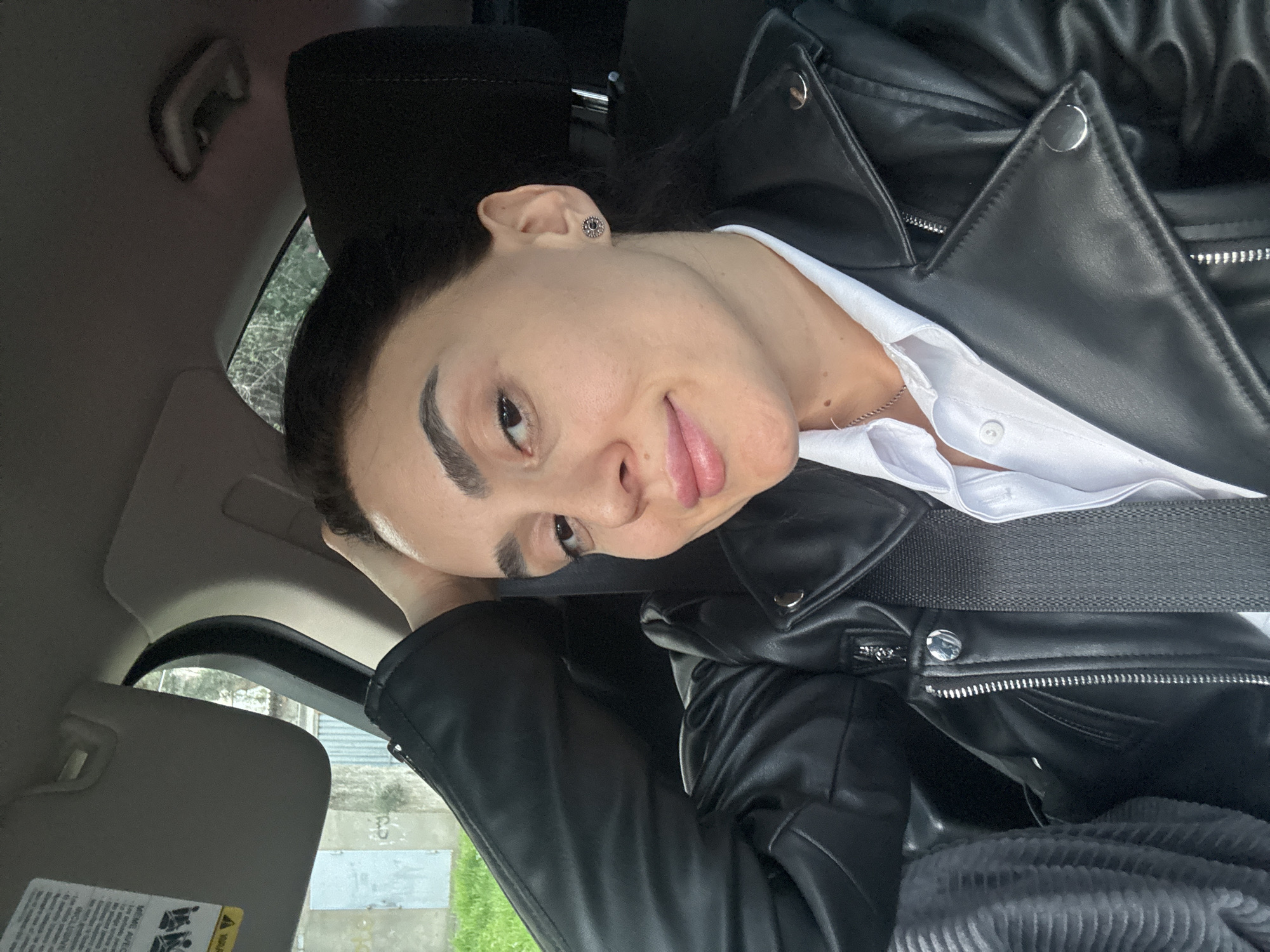 A person wearing a black leather jacket and a white collared shirt is seated in a car. The person is leaning their head against the headrest. The interior of the car is visible, including the sun visor and headrest. A seatbelt crosses the person's chest. Outside the car window, a street scene is visible with greenery and buildings. Based on the environment, it is plausible that this photograph may have been taken in a city environment, such as Tbilisi, Georgia.