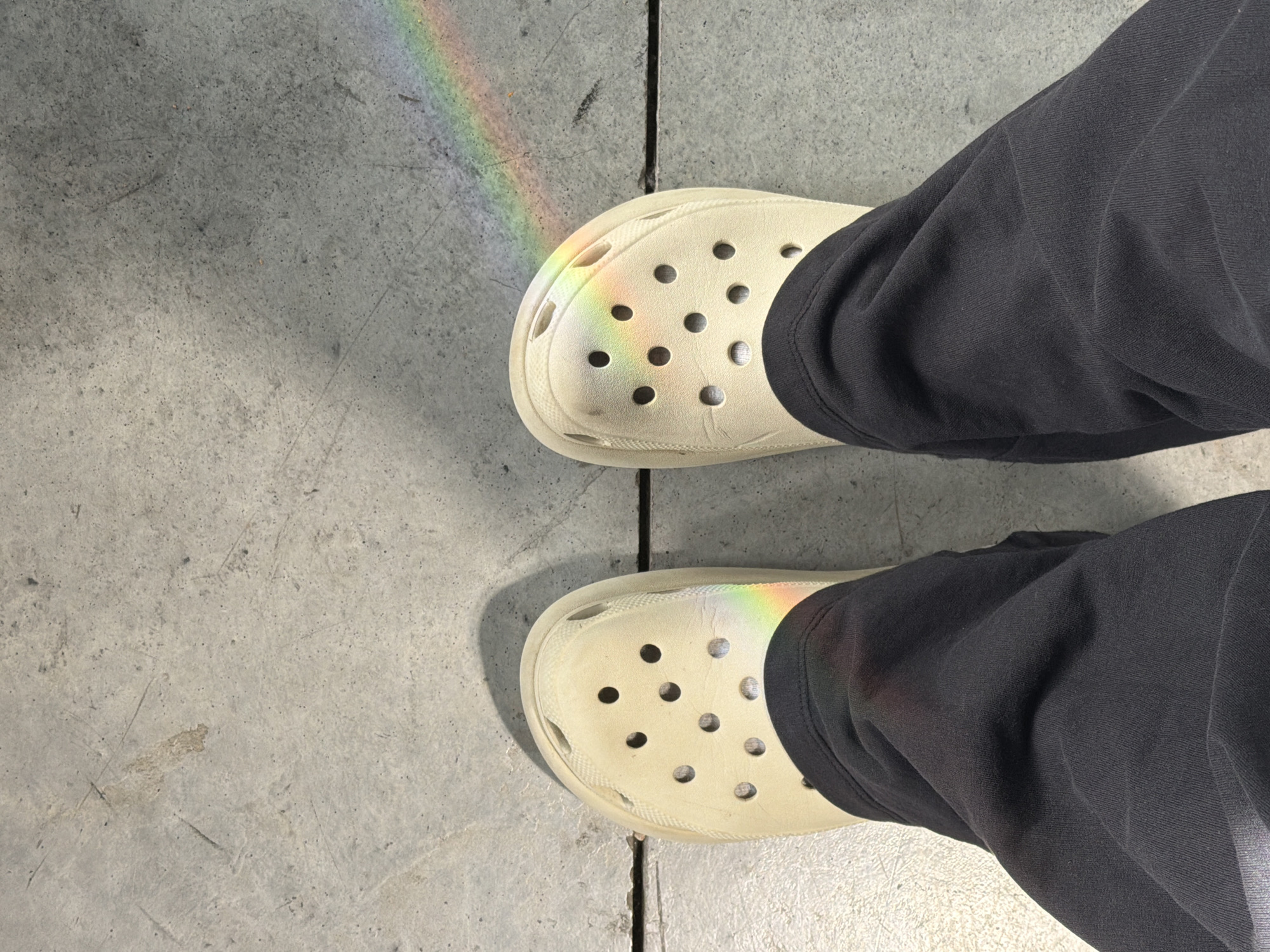 The image shows a person's feet from an overhead perspective. The person is wearing black pants and off-white Crocs. A rainbow is shining across the left side of both shoes and extends onto the gray concrete floor. The floor is divided by dark vertical lines. The location is unspecified, but the scene does not indicate any location in Tel Aviv, Israel.