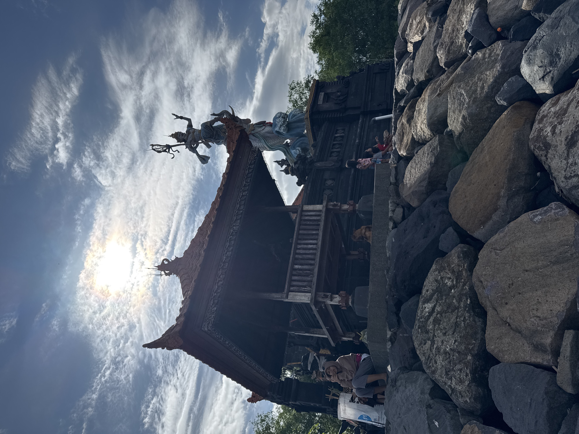 This image depicts a vibrant cultural site under a bright, sunny sky with scattered, wispy clouds. Dominating the upper center is an ornate statue of a mythical figure, possibly a deity or warrior, rendered in muted tones with blue accents, standing atop a traditional Balinese-style pavilion. The figure appears multi-armed, holding a staff or weapon, and is adorned with an elaborate headpiece.
The statue rests on a dark-wood, multi-tiered pavilion, characteristic of Balinese architecture, featuring intricate carvings and a prominent, angled roof. To the right, a substantial wall of large, dark grey rocks forms a natural barrier, extending into the foreground.
Several people are present, visiting or resting at the site. Two children, one in a red shirt and another in a patterned top, are seen resting or lying on a raised stone platform near the base of the pavilion. Closer to the foreground and to the left of the rocks, an adult wearing a light-colored jacket and grey pants is seated amongst the rocks, looking towards the camera. Other less distinct figures are visible near the structure's base.
The scene takes place during bright daylight, with the sun prominently visible in the upper left, creating a strong lens flare and silhouetting parts of the structure. The weather is clear with some scattered white clouds in the blue sky. Green foliage can be seen in the background, suggesting a natural environment. The architecture and statue strongly indicate a location with Balinese cultural influence, consistent with Kediri, Indonesia. A white bag near the seated adult has partial text visible: "ER RESORT".