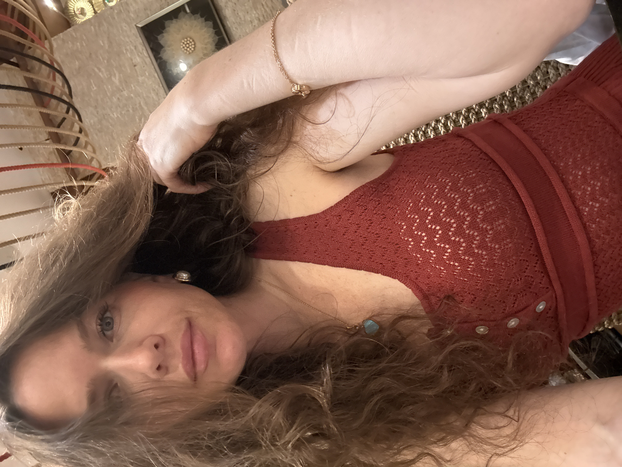 A young adult female is captured in a close-up, front-facing selfie, located indoors in Dubai, United Arab Emirates. She has fair skin, blue eyes, and long, voluminous, curly auburn-brown hair, with strands partially obscuring the left side of her face. Her expression is neutral with a slight hint of a smile.
She is wearing a sleeveless, rust-red knit top or dress featuring an open lace-like pattern, a V-neckline, and a placket with three small, light-colored buttons. Accessories include a gold-toned hoop earring with a spherical pearl-like element on her left ear, a delicate gold-toned chain necklace with a heart-shaped, teal-colored stone pendant, and a thin gold-toned chain bracelet with a small, indistinct charm on her right wrist. Her right hand is raised, with fingers gently touching or running through her hair near her temple.
The background indicates an interior setting with warm lighting. Directly behind her head, a series of thick, colored ropes or cords (beige, red, black) hang vertically from an unseen structure, suggesting a decorative ceiling or wall installation. To her upper right, a light-colored, textured wall is visible, on which a framed piece of art hangs. This artwork displays a large, stylized, light yellow or white circular form with a central cluster of small, pearl-like beads, resembling a dandelion seed head, set against a dark background within a dark frame. Other subtle, metallic-toned decorative elements are faintly visible further in the background.