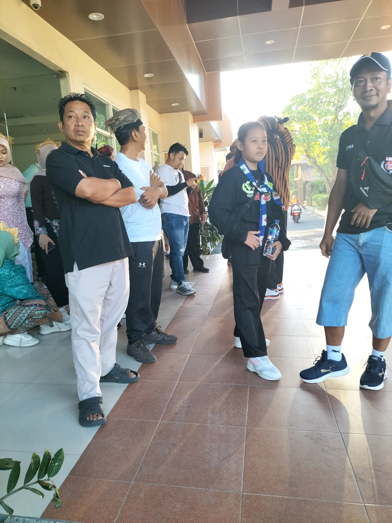 The image depicts an outdoor gathering in front of a building with a tiled walkway. The scene appears to be in Pare, Indonesia, suggested by the context of the surrounding area. In the foreground, a man wearing a black polo shirt, light-colored pants, and dark sandals stands with his arms crossed. To his right, a young person in a black outfit with a colorful scarf holds a bottle of water. Further to the right, a man in a black shirt and blue shorts, wearing a baseball cap, is visible. Several other individuals are partially visible in the background, including people in traditional attire. The environment includes architectural elements of the building and some greenery. The lighting suggests it is daytime.