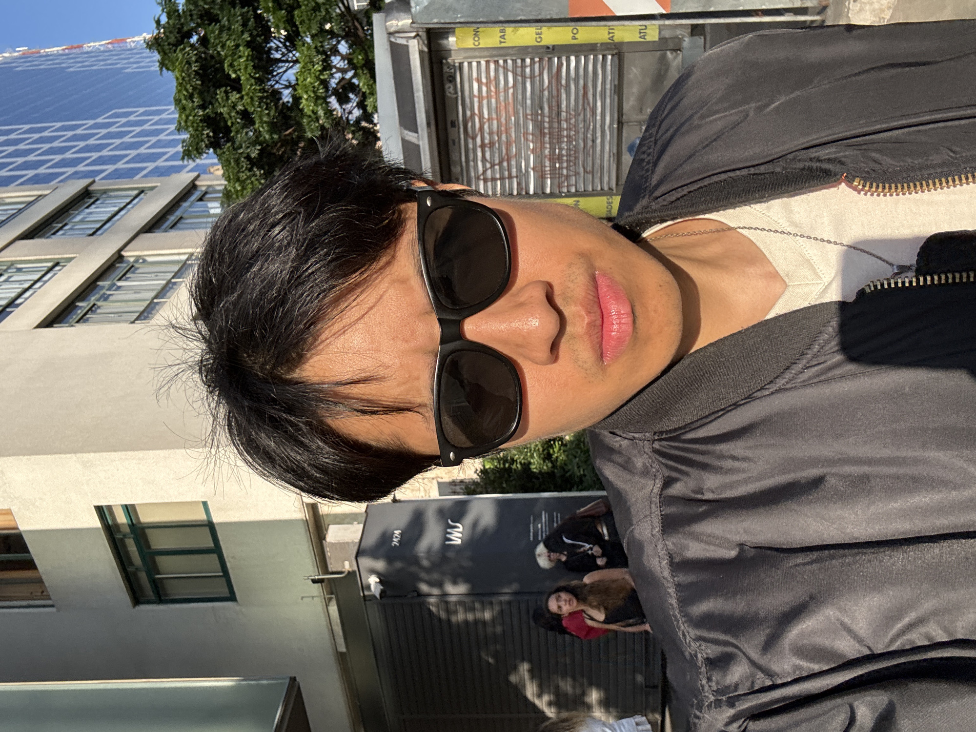 A medium shot presents a person wearing sunglasses and a jacket. Buildings and trees are visible in the background. Other individuals can be seen in the background. The image was taken outdoors. Based on the signage "VAS", the location is likely in São Paulo, Brazil.