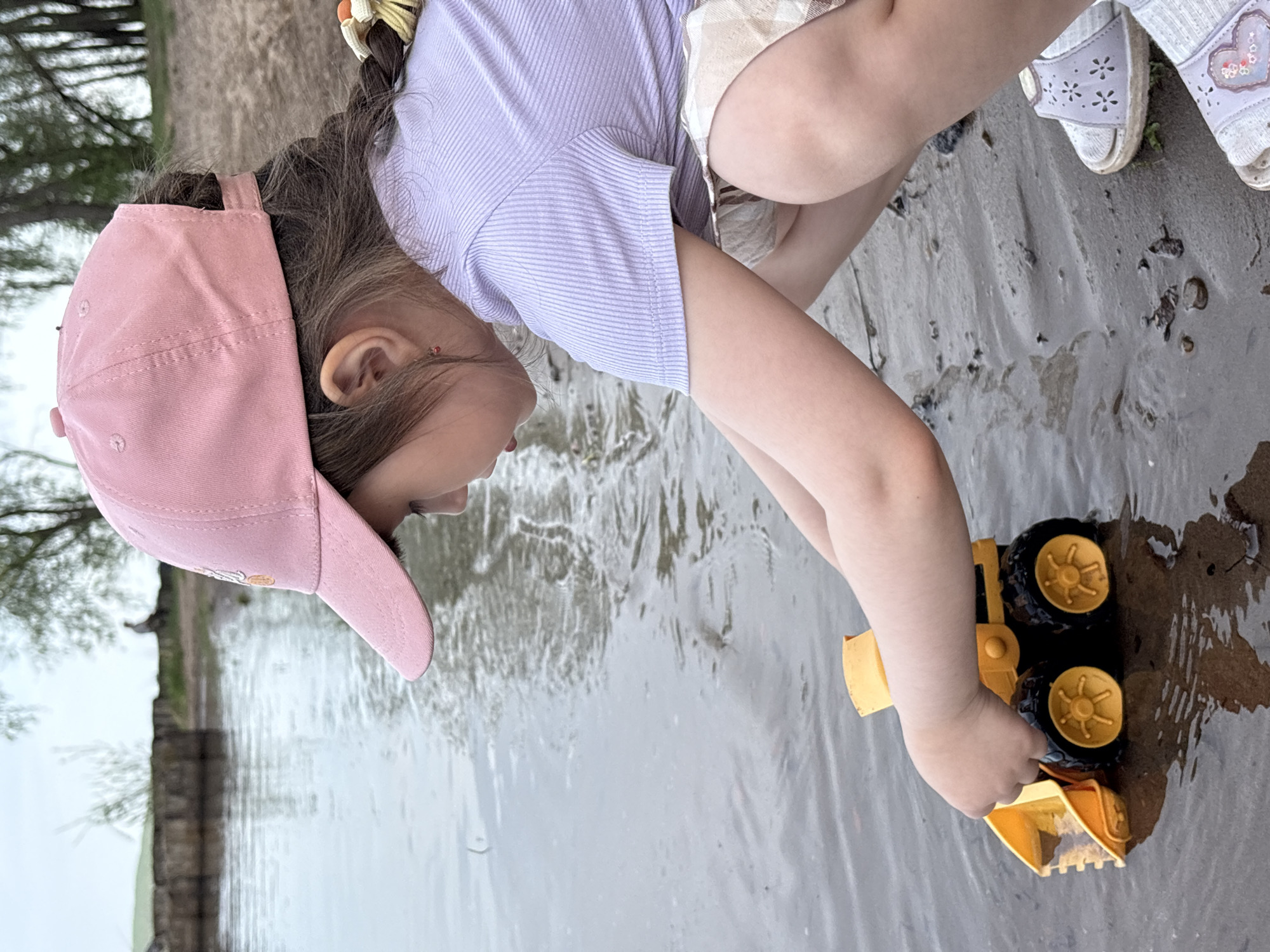 A young child wearing a light purple shirt, patterned shorts, and lilac sandals with decorative hearts and flowers plays with a yellow and black toy bulldozer in the water. The child is wearing a pink baseball cap and has a braid with a yellow and orange hair tie. The location appears to be near a body of water with a shoreline. The image shows a treeline in the background with some structures possibly indicating it is in the city of Chaikovsky, Russia.