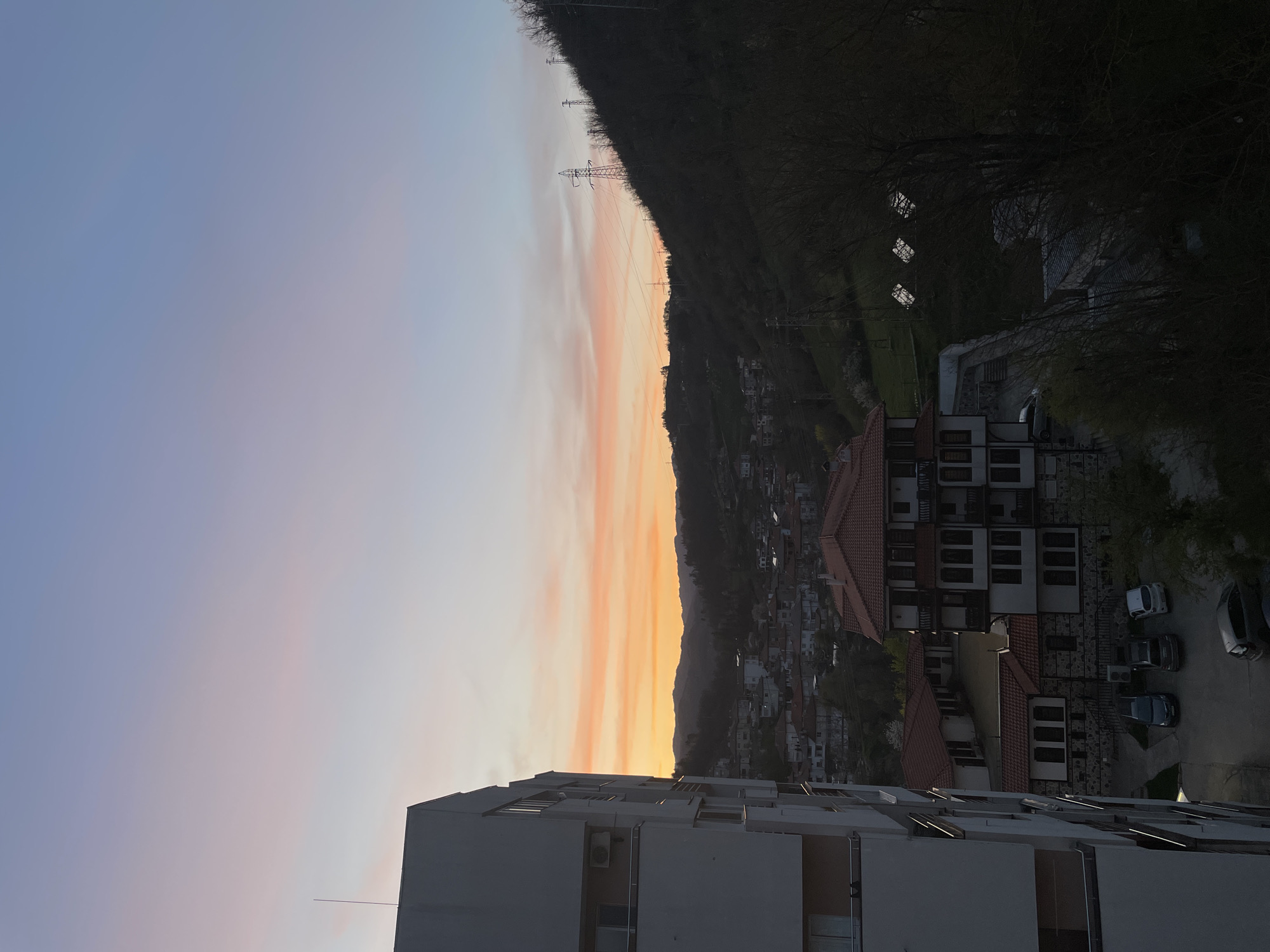 The image depicts a cityscape at sunset, possibly in Zlatograd, Bulgaria. The sky transitions from blue to orange hues. A mountain range is visible, partially obscured by trees and with electrical pylons. Buildings with terracotta roofs are clustered together. Cars are parked in a lot near some of the buildings. A multi-story building occupies the foreground.