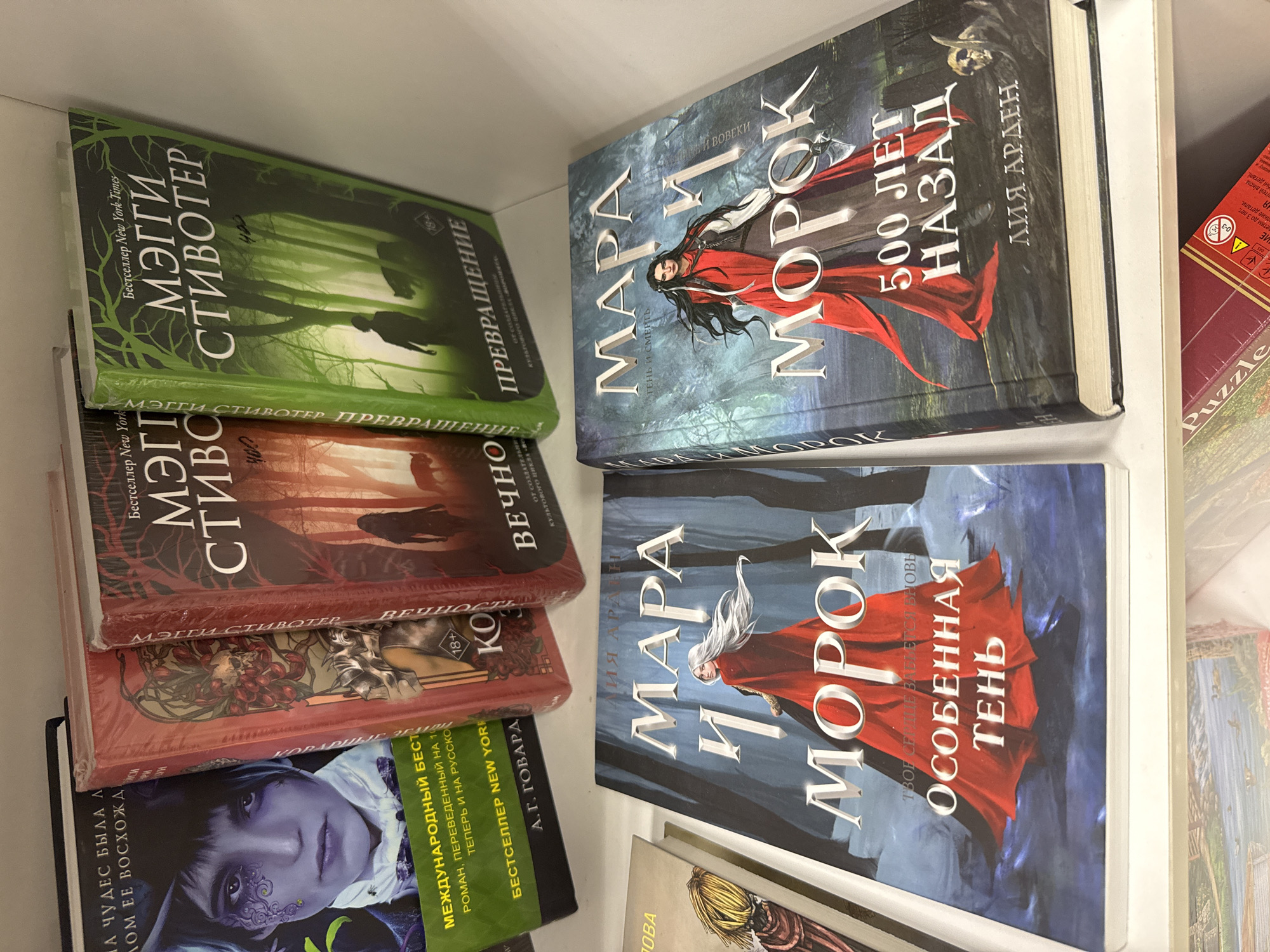 The image shows a shelf with several books, primarily fantasy novels. In the top row, there are at least three books with dark covers, two of which appear to be part of a series based on the repeating title "МЭГГИ СТИВОТЕР". The leftmost book in the top row has a close-up of a person's face with dramatic makeup. Below these, on the bottom shelf, are two larger books with similar cover art featuring women in red cloaks, with titles including "МАРА И МОРОК". The overall impression is of a collection of fantasy literature. The location is identified as Votkinsk, Russia, but there are no specific visual cues in the image to confirm this location, such as landmarks or signs in Russian that would uniquely identify it beyond the text on the books. The environment appears to be an indoor setting, likely a bookstore or a home library, with white shelving. There are no people visible in the image, nor any actions or interactions taking place. The objects are static and arranged for display.