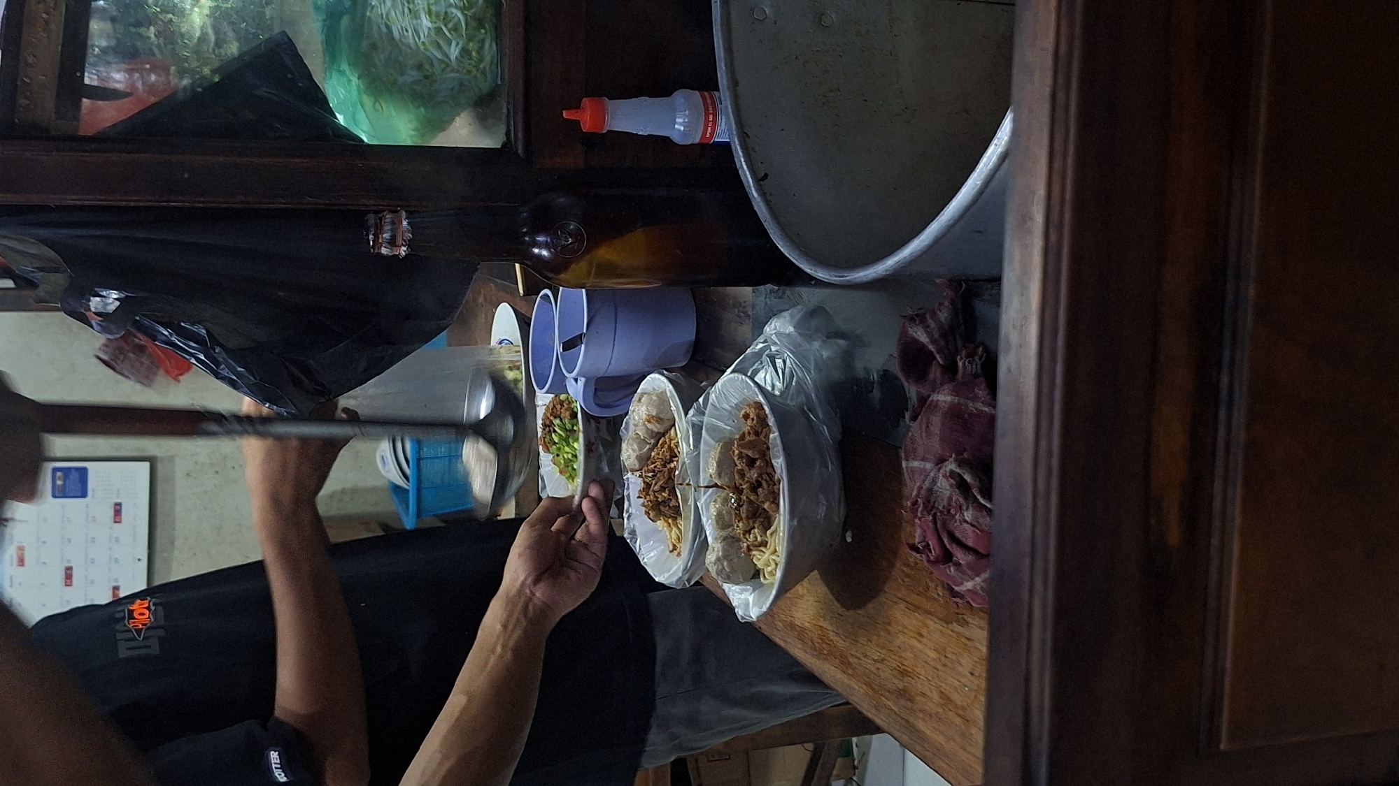 The image shows a street food vendor in Sukoharjo, Indonesia, preparing a meal. A person wearing a black shirt is holding a ladle in one hand and reaching for a bowl of food with the other. There are three bowls containing noodles, meat, and vegetables on a wooden surface. To the left are purple mugs, and in the background, there's a large pot, condiments, and ingredients storage, suggesting a makeshift kitchen setup within a stall or cart. A calendar is visible behind the person preparing the food.