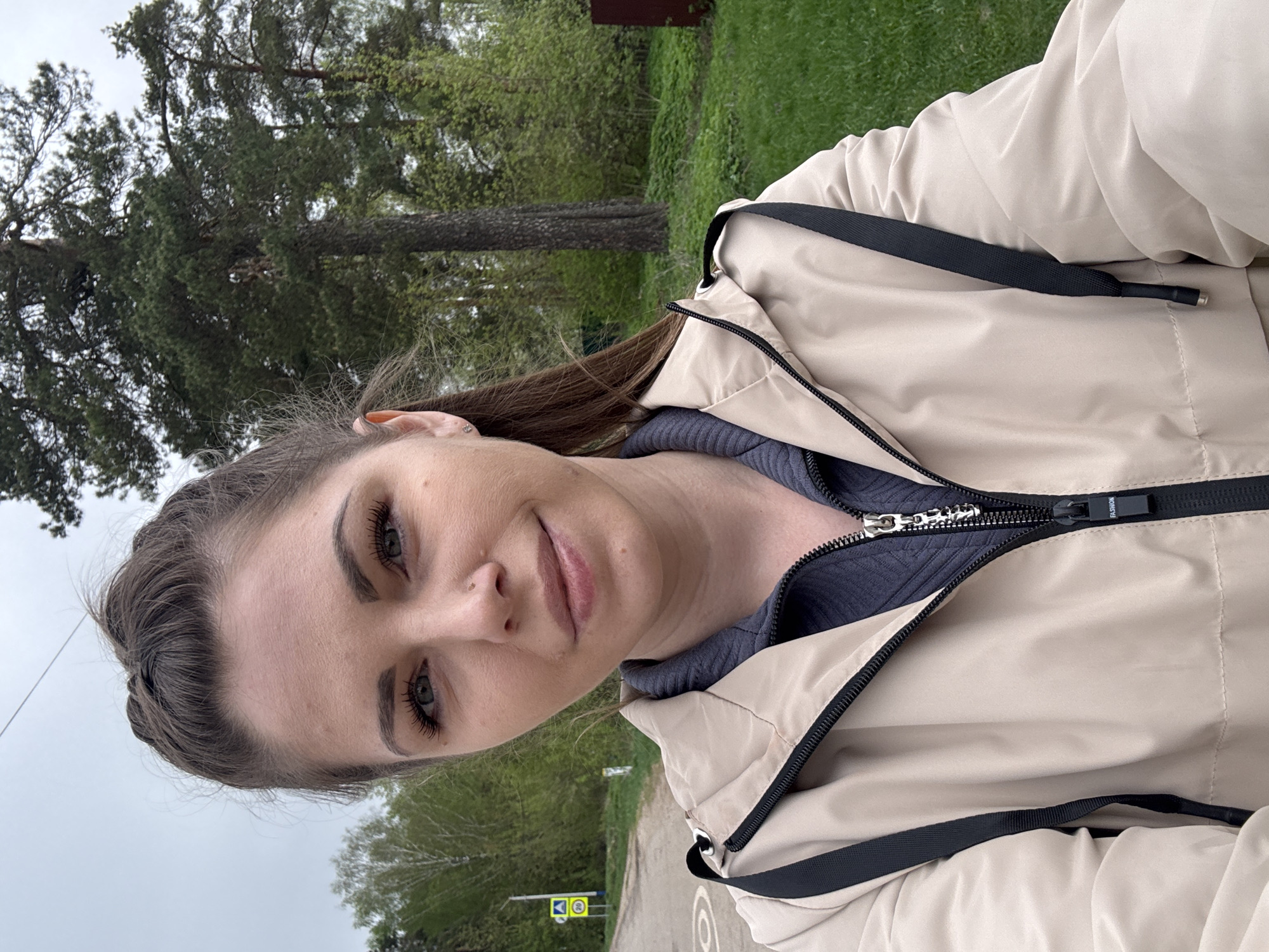 A woman is centered in the image, wearing a beige jacket with black trim, partially zipped over a gray top. The background reveals an outdoor setting in what appears to be Раздолье, Russia, indicated by a yellow road sign with Cyrillic text. Trees are visible in the background.