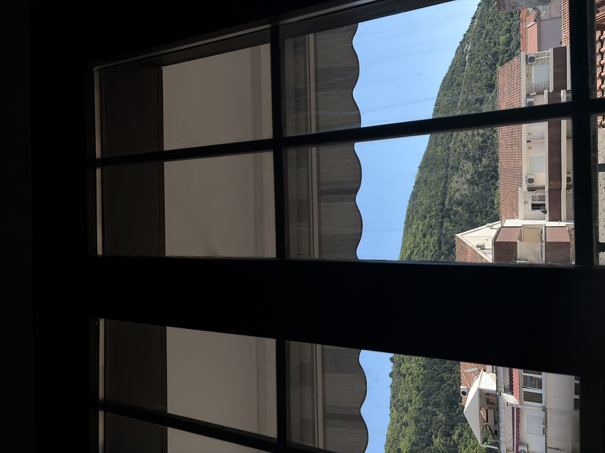 The image is presented rotated 90 degrees clockwise, capturing a view from an interior space looking outwards through a window. A dark, prominent window frame, possibly made of wood or metal, divides the scene with horizontal and vertical mullions, creating a grid of panes.
On the left side of the rotated image (which would be the top when viewed upright), the view is largely of a dimly lit interior, showing parts of a wall or curtain in muted grey and white tones.
The right side of the rotated image (the bottom when viewed upright) reveals a bright exterior scene under a clear, light blue sky. A large, densely forested green mountain dominates the background, rising steeply and stretching across the frame. In the mid-ground, several multi-story buildings with distinct red-tiled roofs are visible. These structures feature numerous windows, balconies, and external air conditioning units mounted on their facades. A striped awning with a decorative scalloped edge is partially seen through one of the upper windowpanes, adding a touch of color.
The scene is captured during bright daylight with clear weather, indicating a sunny day. No people are present, and there is no visible text. The architectural style and mountainous coastal landscape are consistent with a location like Budva, Montenegro, suggesting a scenic view from a room overlooking the town and its natural surroundings.