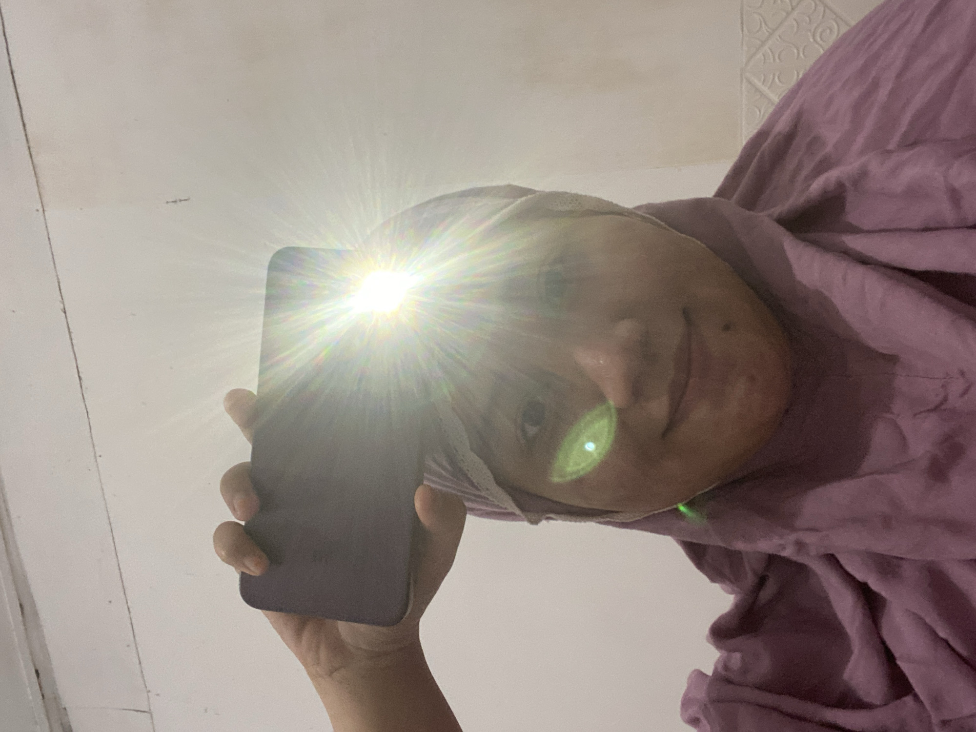 The image shows a close-up of a person's face and upper body, taken from a slightly low angle. The person is wearing a purple hijab and a purple garment that appears to be a hijab or a loose-fitting top. The person is holding a smartphone horizontally above their head with their right hand, and the phone's flash is on, creating a bright glare that obscures the top of the face and emits light rays. The background is a plain, light-colored wall with some subtle texture or pattern at the bottom right. There is a green lens flare on the person's cheek. The overall lighting is harsh due to the direct flash. The image appears to be a selfie taken indoors. There is no visible indication of the specific location in Banjarbaru, Indonesia, beyond it being an indoor setting.