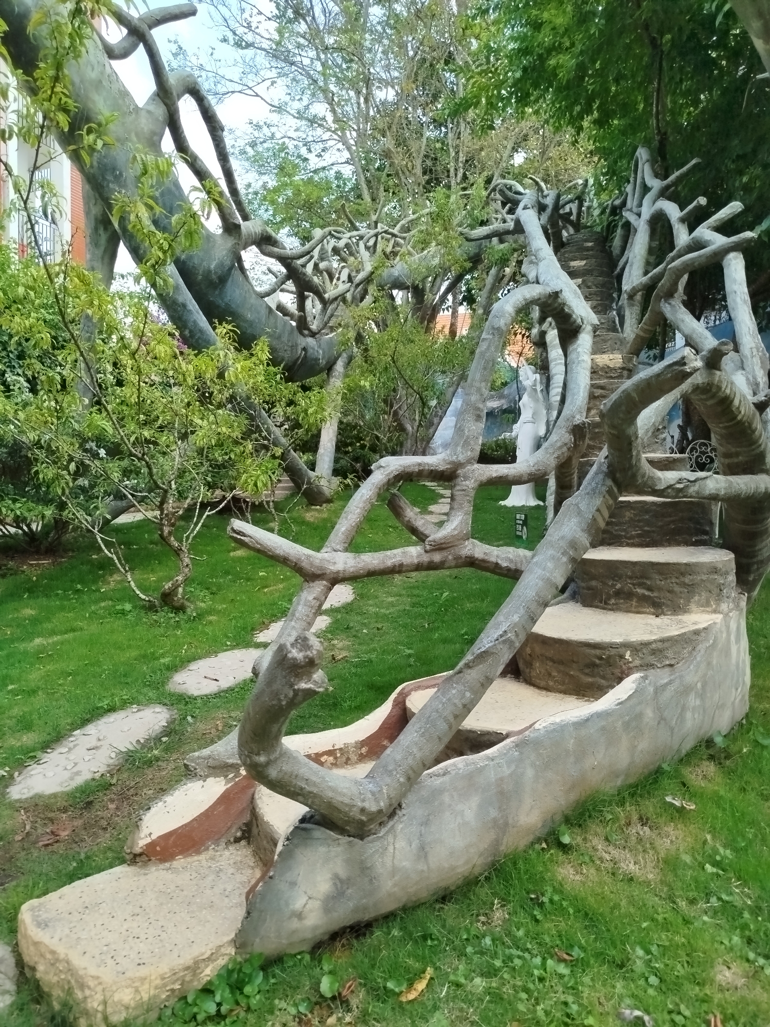 The image depicts an unconventional staircase structure in Da Lat, Vietnam, characterized by its design resembling intertwined tree branches. The staircase is constructed from concrete, shaped to mimic the appearance of a natural tree, with the steps integrated into the framework of the “branches.” The staircase is situated in a lush, green garden environment with interspersed patches of grass and stone pathways. A few leafy bushes and trees are also visible in the scene. In the background, a partial view of a building with white walls is present. A white statue is partially visible in the middle ground.