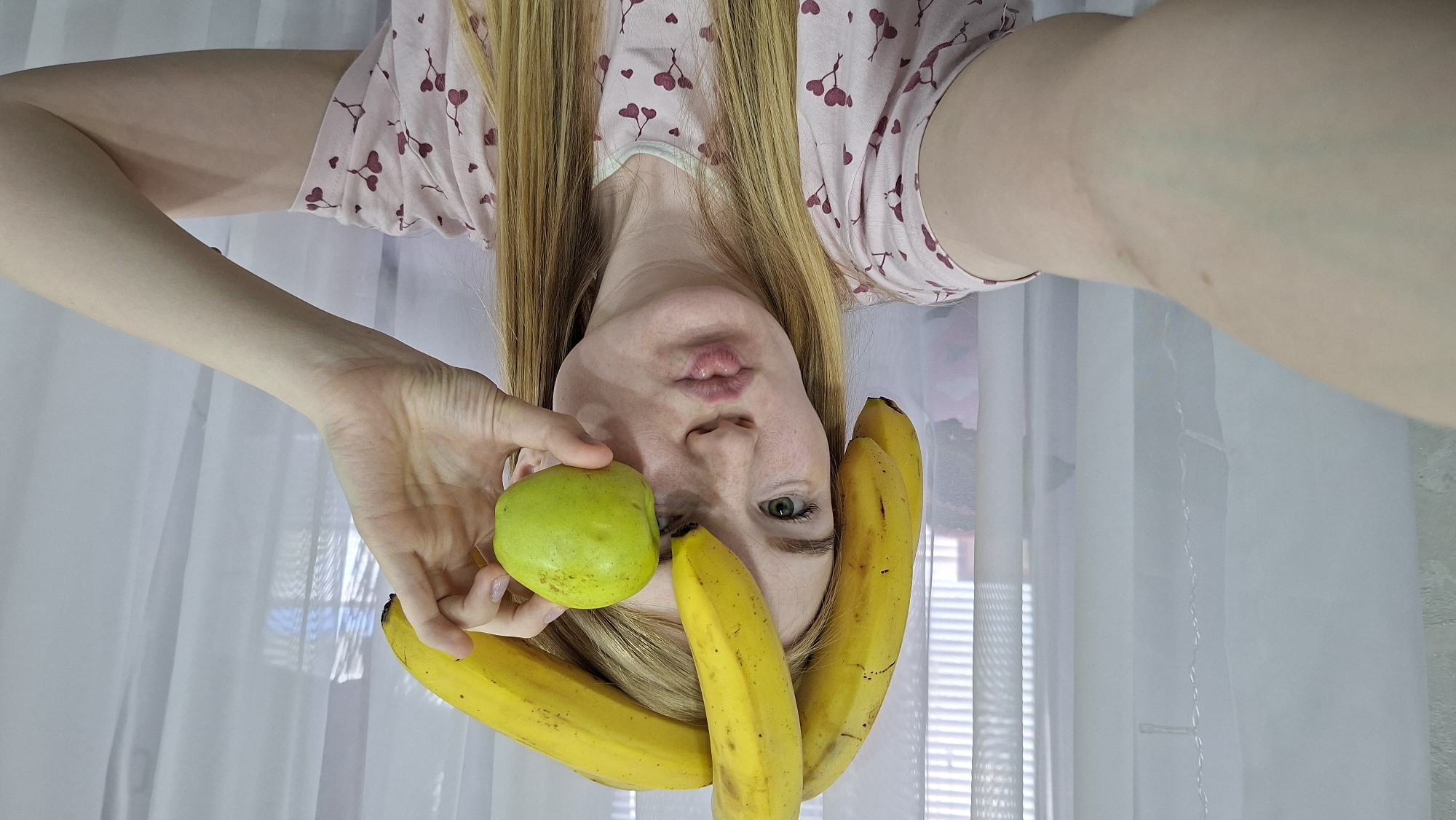The inverted image shows a young woman with long, straight blonde hair, fair skin, and green eyes, appearing to be suspended upside down. She is wearing a light-colored t-shirt patterned with small, dark heart shapes. Her mouth is puckered in a playful expression, and her gaze is directed towards the camera.
In her left hand, she holds a bright green apple close to her face. Three yellow bananas are arranged on and around her forehead, with one seemingly nestled into her hair or held by her right hand, which is mostly out of view.
The background consists of translucent white sheer curtains, through which the slats of window blinds are faintly visible, suggesting an indoor setting during daylight hours. The soft, natural light illuminates the scene. The overall impression is one of a lighthearted or humorous moment. No visible text is present. The scene is set in Chaikovsky, Russia.
