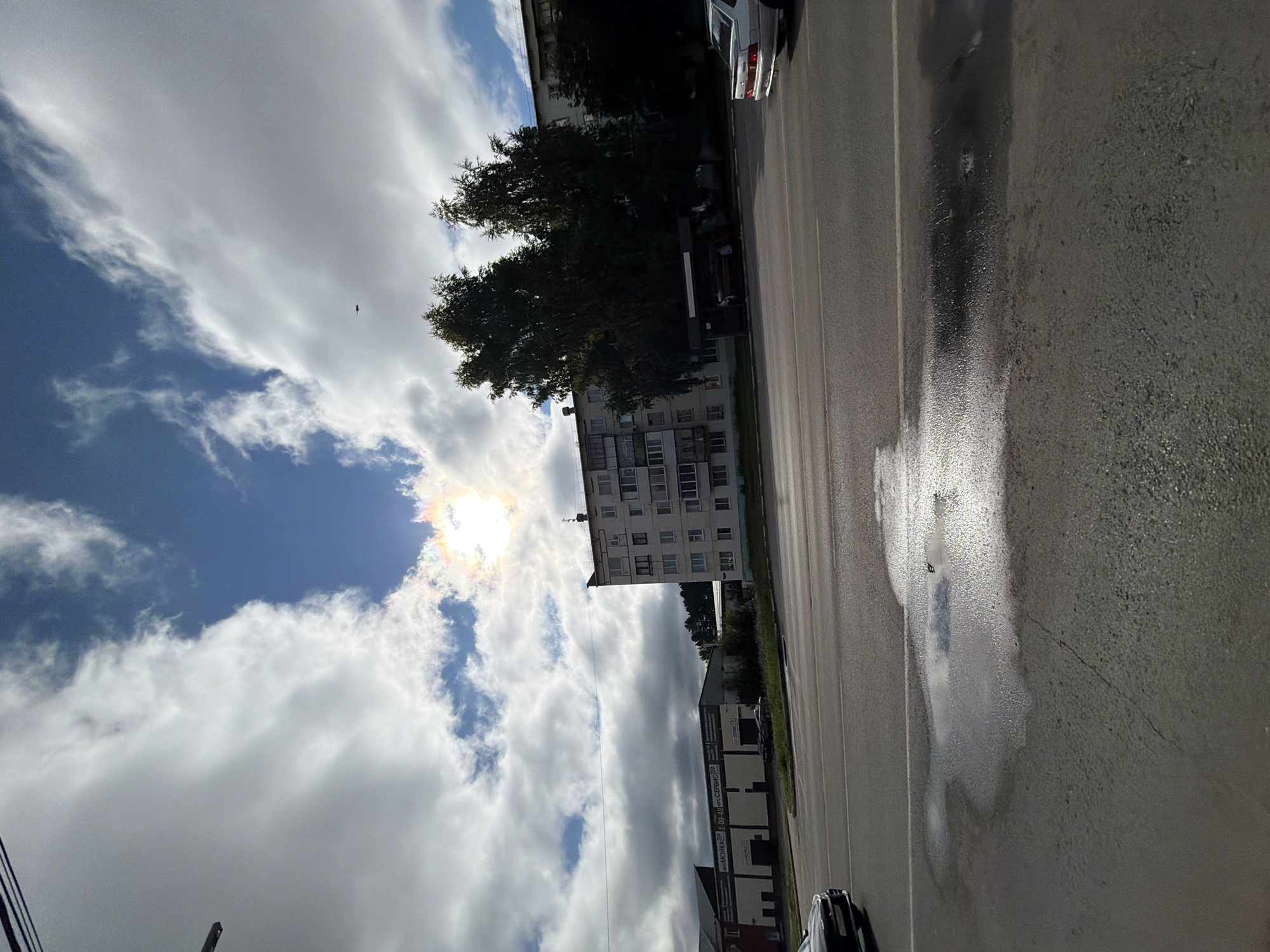 The image depicts a street scene in Chaikovsky, Russia, on a partially cloudy day. In the foreground, a wet asphalt road is visible, with a large puddle reflecting the sky. A dark-colored car is partially visible on the left edge of the frame, driving on the road. In the middle ground, across the road, there is a row of low-rise buildings, one of which appears to be a commercial establishment with signage. To the right of these buildings, there are several tall, mature trees. Further to the right, a light-colored sedan is parked. The background is dominated by a bright sky with a mix of white clouds and patches of blue. The sun is visible, partially obscured by clouds, creating a halo effect. A small dark object, possibly a bird or drone, is discernible in the sky. The overall impression is that of an urban environment on a day that may have recently experienced rain.