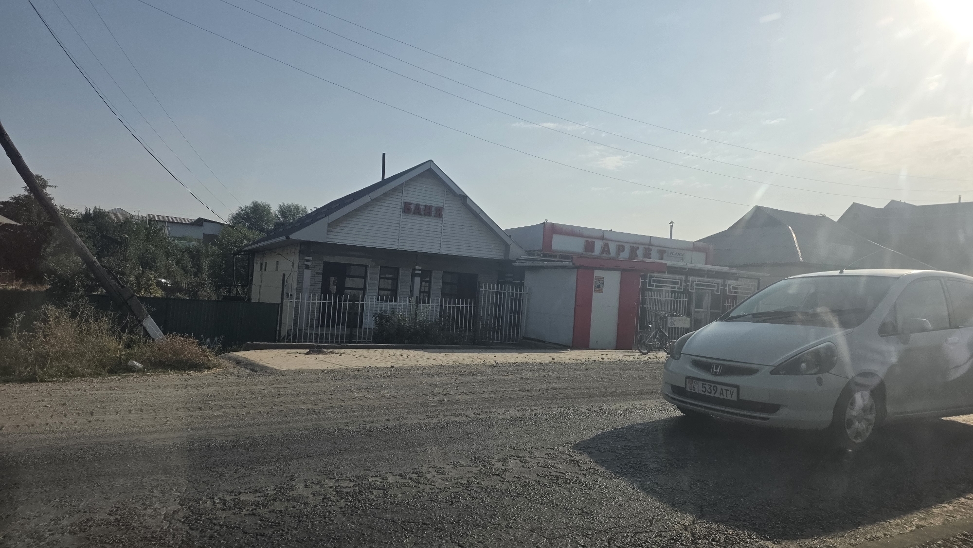 The image depicts a street scene in the city of Baş-Bulak, Kyrgyzstan. On the right side of the street, a light-colored Honda Fit car, license plate "06 539 ATY", is parked. In the background, there is a building complex with a white, gabled structure that has the word "БАНЯ" (Banya) in red lettering across its facade. Adjacent to this building is another structure with red and white signage that reads "МАРКЕТ" (Market). A bicycle is parked in front of the market. The buildings appear to be single-story commercial establishments. The environment is rural or semi-urban, with a dirt road in the foreground and trees and other buildings visible in the distance. Overhead, power lines are strung across the sky. The lighting suggests it is daytime, with a clear sky and sunlight creating some lens flare.