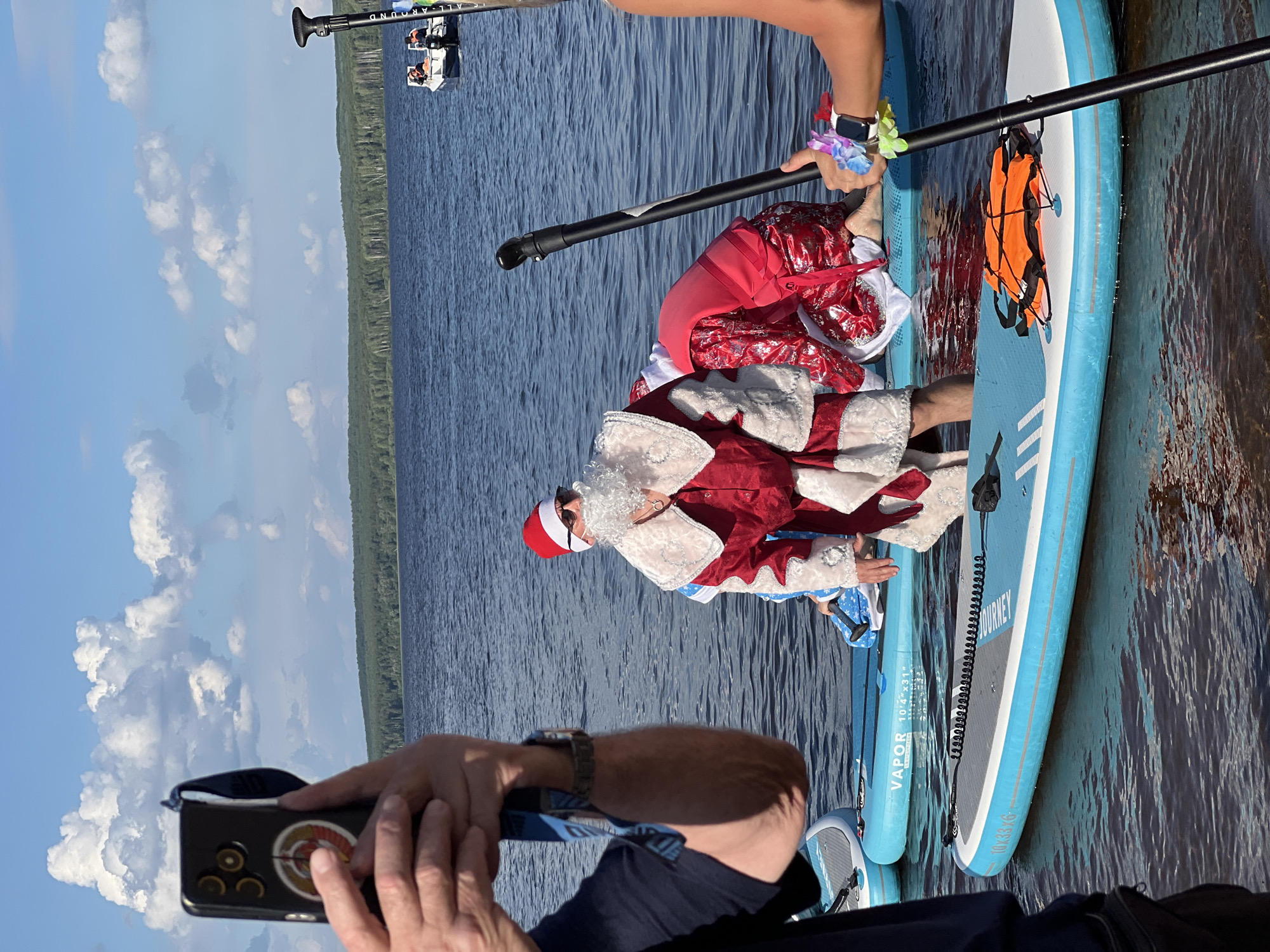 In this image, two people are on paddleboards in a body of water. The person in the foreground, wearing a blue shirt and a watch, is holding a smartphone and appears to be taking a photo. The person on the paddleboard behind them is dressed as Santa Claus, complete with a red suit, white beard, and hat. They are holding a paddle and are partially leaning over their paddleboard.
The scene is set on a lake or large body of water, with a tree-lined shore visible in the background. The sky is blue with scattered white clouds, suggesting it is daytime with good weather. There are other paddleboards and some smaller boats further in the distance, indicating a possible recreational event or gathering.
On the paddleboard with Santa, there are two orange life vests clipped to the side. Text is visible on the paddleboard itself, including "VAPOR 10'4" x 31"" and "JOURNEY 10'4" x 33" x 6"". The overall mood appears festive and relaxed, with the Santa costume adding a touch of holiday spirit to the outdoor activity. The image appears to be taken in Chaikovsky, Russia, given the location context.