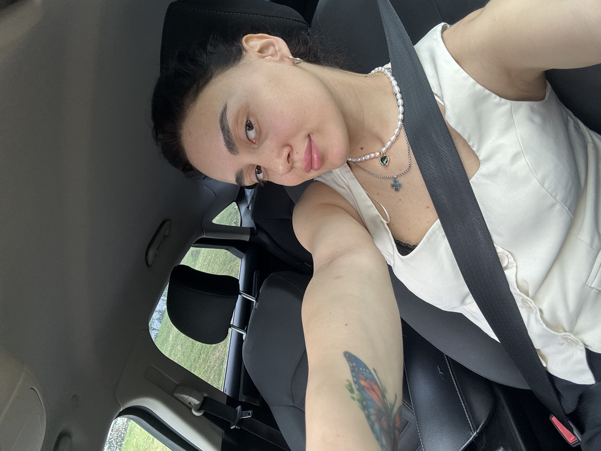 A fair-skinned woman is seated in a car, wearing a white sleeveless top, a pearl necklace with a heart and cross pendant, and a seatbelt. Her left arm, featuring a butterfly tattoo, is extended towards the camera. The interior of the car includes black leather seats and a view of the exterior through a window with raindrops. The location, Tbilisi, Georgia, cannot be definitively determined from the provided visual information.