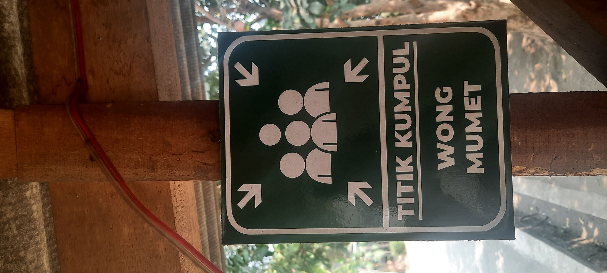 A green sign with white text and symbols is mounted on a wooden post in Ploso, Indonesia. The sign displays an icon of three stylized people figures with arrows pointing towards them from the top and sides. Below the icon, the text reads "TITIK KUMPUL" and "WONG MUMET". The surrounding environment suggests an outdoor setting with trees and a paved walkway visible in the background. The sign appears to be a designated assembly point, possibly for a gathering or in case of an emergency.