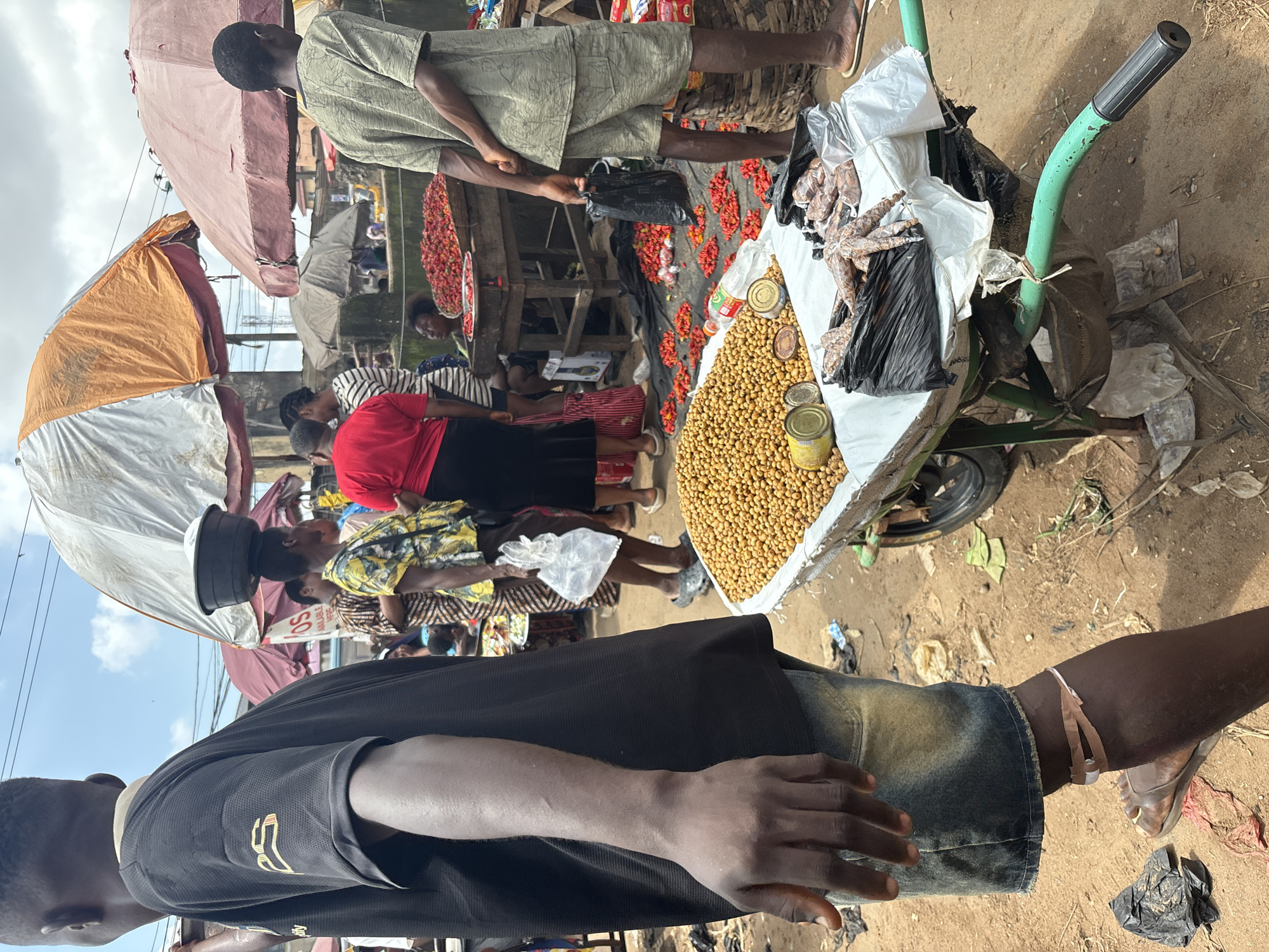 In Fegge, Nigeria, this outdoor market scene depicts a bustling environment. In the foreground, a man wearing a black t-shirt and denim shorts stands to the left, facing away from the camera. His right arm is visible, and his left leg is partially obscured. In front of him, a cart is laden with a large quantity of small, yellow, spherical food items, possibly beans or nuts, along with several cans. Bags of what appear to be dried fish or meat are also present on the cart.
Behind the man, other market-goers are seen. A woman in a yellow and black patterned shirt carries a black bowl on her head. Another woman wears a red top and a dark skirt, and further back, a person in a striped shirt is visible. Various market stalls with umbrellas provide shade. Piles of red peppers are displayed on the ground and on wooden tables, indicating a lively commercial exchange of produce. The ground is dirt and appears somewhat dusty, with scattered debris. The overall atmosphere suggests a typical day at a busy Nigerian marketplace.