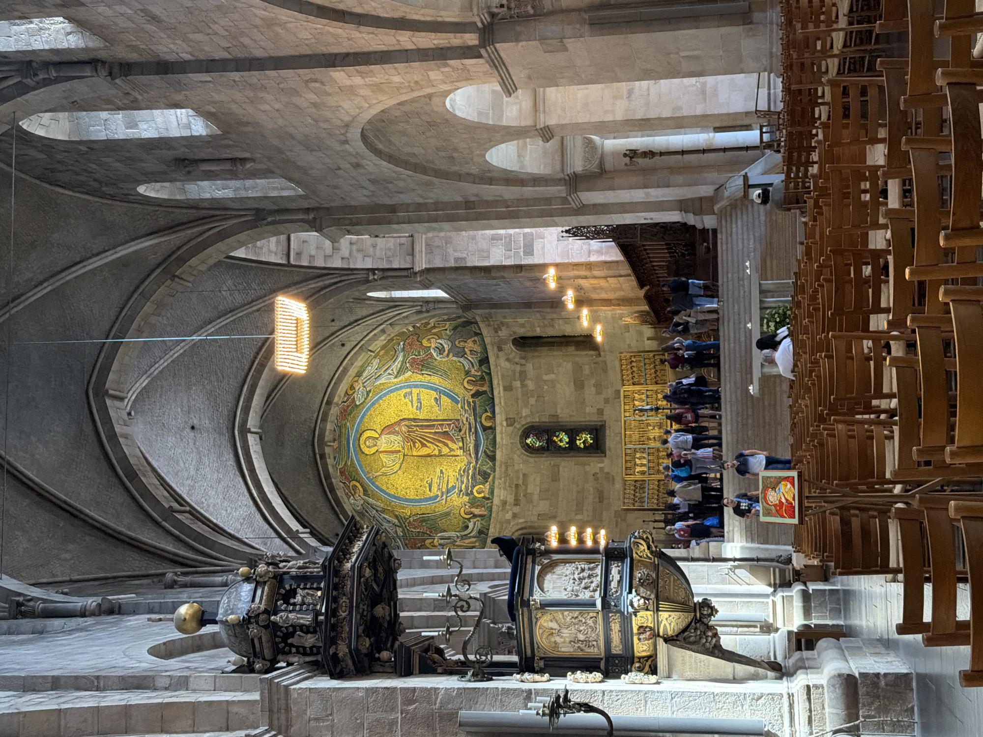 The image shows the interior of Lund Cathedral in Lund, Sweden. The cathedral features a large, circular mosaic above the altar depicting Christ. In the foreground, rows of wooden chairs fill the nave. A pulpit, ornately carved and decorated, stands to the left of the frame. Several people are visible throughout the cathedral, some standing in groups, others seated in the chairs, and a few walking. Natural light streams in through arched windows high on the walls, illuminating the stone architecture and the golden mosaic. The overall impression is one of historical grandeur and serene atmosphere.
