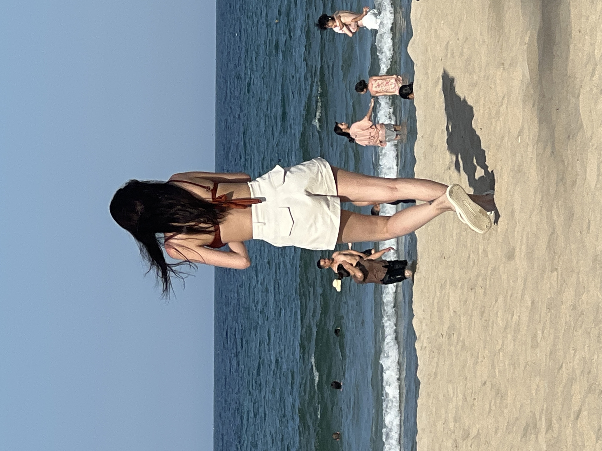 In Đà Nẵng, Vietnam, on a sun-drenched beach, a woman with long, dark hair, viewed from behind, stands on the sand. She is wearing a reddish-brown bikini top and high-waisted, cream-colored shorts with prominent pockets. Her hair is windblown, and she is wearing simple white flip-flops. She is facing the ocean, which is a vibrant blue with moderate waves breaking near the shore.
Several other people are present in the water. In the mid-ground, a man in a brown short-sleeved shirt and dark shorts is partially submerged, looking towards the woman. To his right, a person in a pink shirt and grey shorts is interacting with a child wearing a patterned dress. Further to the right, a person holding a child stands in the water. Other individuals are visible as heads or torsos in the water, indicating various people are enjoying the ocean. The beach itself is composed of light-colored sand, with a shadow cast by the woman in the foreground onto the sand. The sky is a clear, pale blue, indicating a bright and clear day.