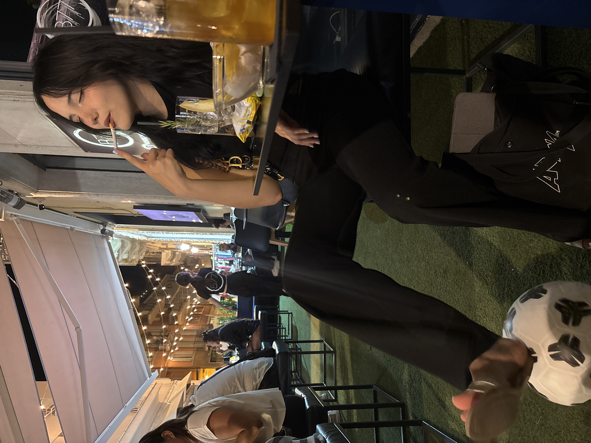 A young woman with long, dark hair sits at a table outside a bar in San Benedetto del Tronto, Italy, with a cigarette in her mouth and her eyes closed. She is wearing a black top and black pants, and her right leg is extended, with her foot resting on a soccer ball. A glass with a drink and ice, a sprig of rosemary, and a slice of lemon is on the table in front of her, along with a bag of chips. Behind her, string lights are visible, illuminating the outdoor seating area of the establishment. The scene appears to be at night.