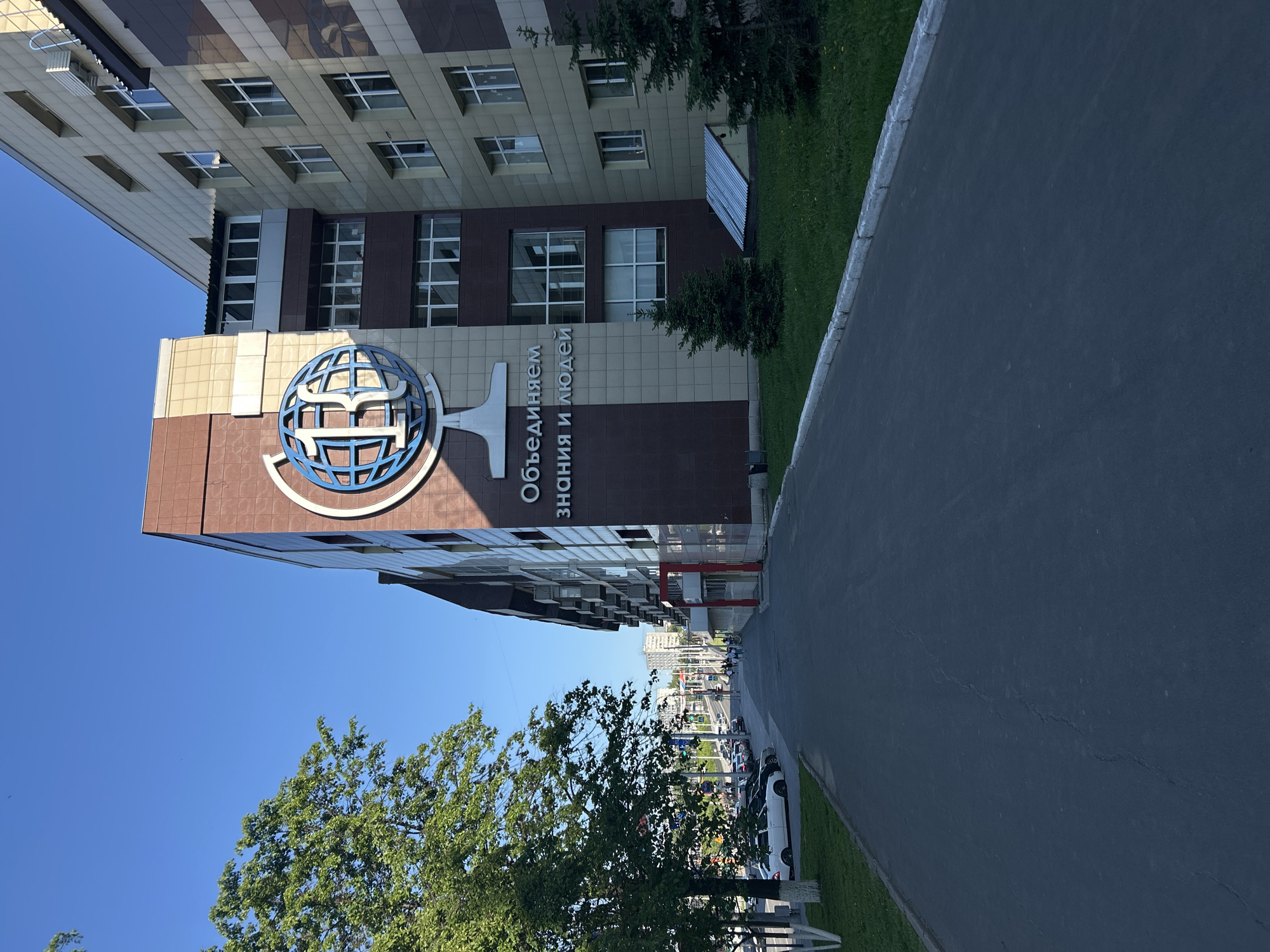 The image captures a modern building under a clear, bright blue sky on what appears to be a sunny day. The building's facade features a distinctive pattern of light beige, brown, and darker brown square tiles, interspersed with numerous rectangular windows reflecting the sky.
A prominent blue and white emblem is affixed to one side of the building, depicting a stylized globe or sphere with interconnecting elements. Adjacent to this emblem, white Russian text is displayed vertically, reading "Объединяем знания и людей," which translates to "Uniting knowledge and people."
To the left foreground, a mature tree with lush green foliage partially obstructs the view of the building's base. On the right side of the frame, an asphalt path or road with a concrete curb runs diagonally, bordered by a strip of green grass and some smaller trees or bushes. In the background, beyond the main building, glimpses of other urban structures and parked vehicles are visible, suggesting an urban or suburban setting.
The scene is static, with no discernible activity or individuals present. The overall impression is one of a clean, well-maintained environment around a contemporary structure, likely an educational, research, or corporate institution given the motto displayed. The image itself is rotated 90 degrees clockwise from its natural orientation.