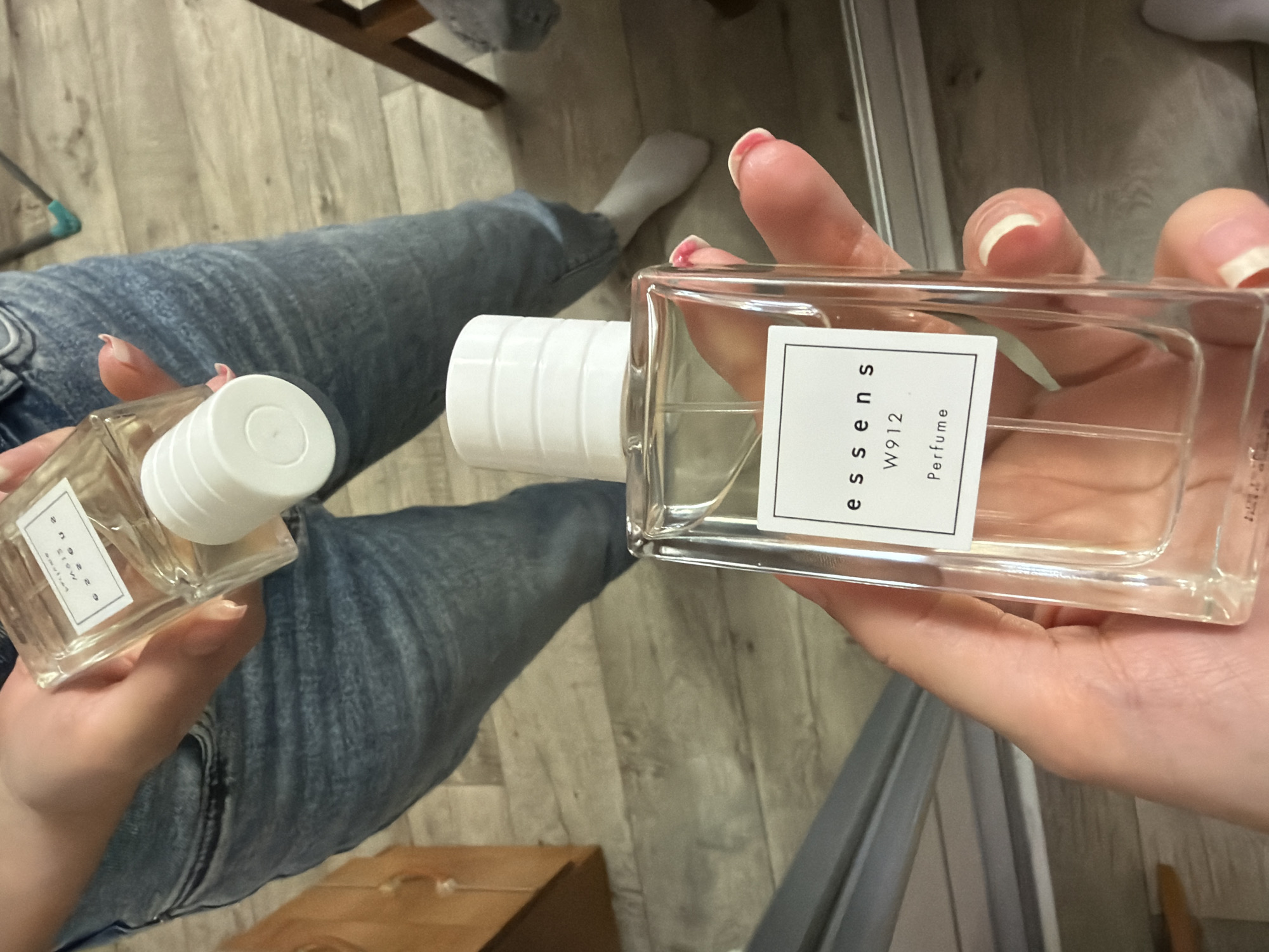 In a casual indoor setting, two perfume bottles are held by unseen individuals. The foreground features a clear glass bottle with a white cap and a white label that reads "essens W912 Perfume". The background shows another perfume bottle, also clear glass with a white cap and a label with text that is difficult to decipher. The individuals are wearing denim jeans. The flooring appears to be made of light-colored wood planks. The background includes a wooden cabinet and part of a chair. There is no information in the image to suggest any specific location within Tuymazy, Russia, or any other city.