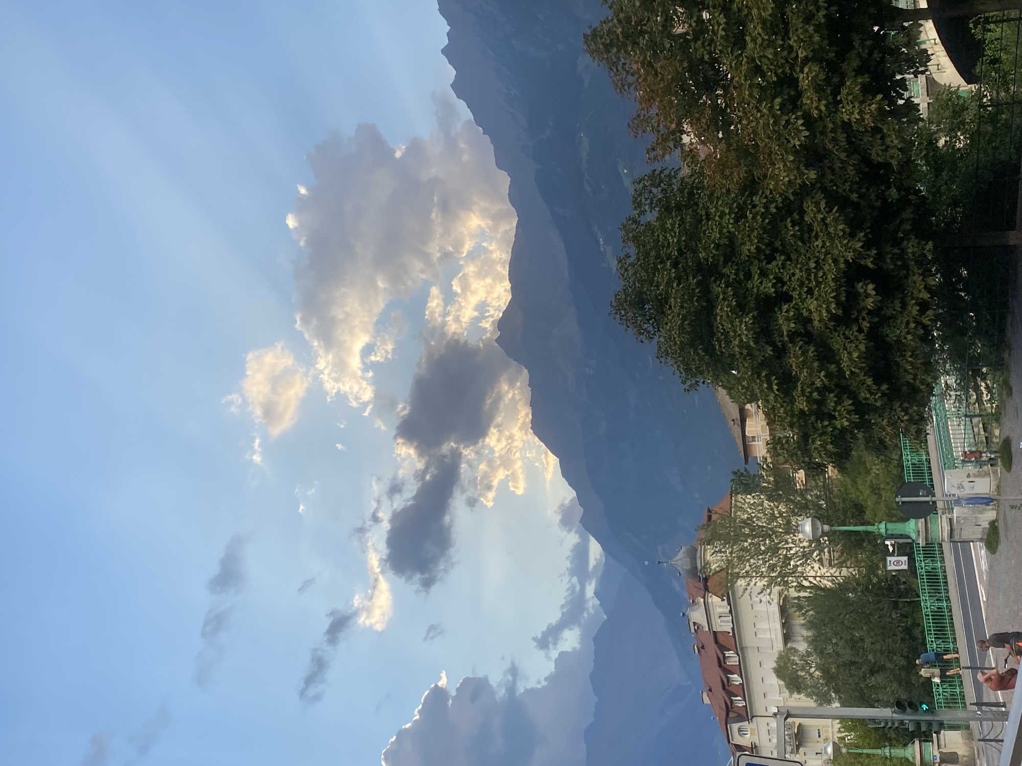 The image captures a view of the city of Meran (Merano), Italy, with a backdrop of imposing mountains. In the foreground, a street scene unfolds, featuring a bright green bridge adorned with decorative lampposts. Two individuals are visible standing on the bridge, and a few more are seated on the sidewalk. Buildings with red-tiled roofs line the street, and lush green trees are scattered throughout the scene. The sky is a pale blue, punctuated by scattered clouds, with rays of sunlight breaking through them, casting a dramatic effect. The overall impression is one of a serene, late afternoon or early evening in a picturesque European city.