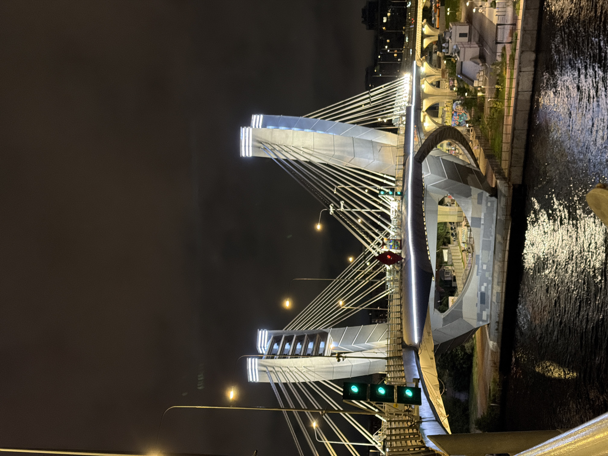 The image shows a nighttime view of the cable-stayed Bolshoy Obukhovsky Bridge in Saint Petersburg, Russia. The bridge is illuminated by white lights on its towers and along the deck. The foreground features a dark body of water reflecting the lights of the bridge and surrounding cityscape. A traffic light with green signals is visible on the left side of the bridge. The bridge's distinctive modern design includes two V-shaped concrete towers supporting the deck with numerous cables. In the background, other bridges and city lights are visible against a dark, cloudy sky. The overall scene is a well-lit, urban nightscape with a focus on the architectural grandeur of the bridge.