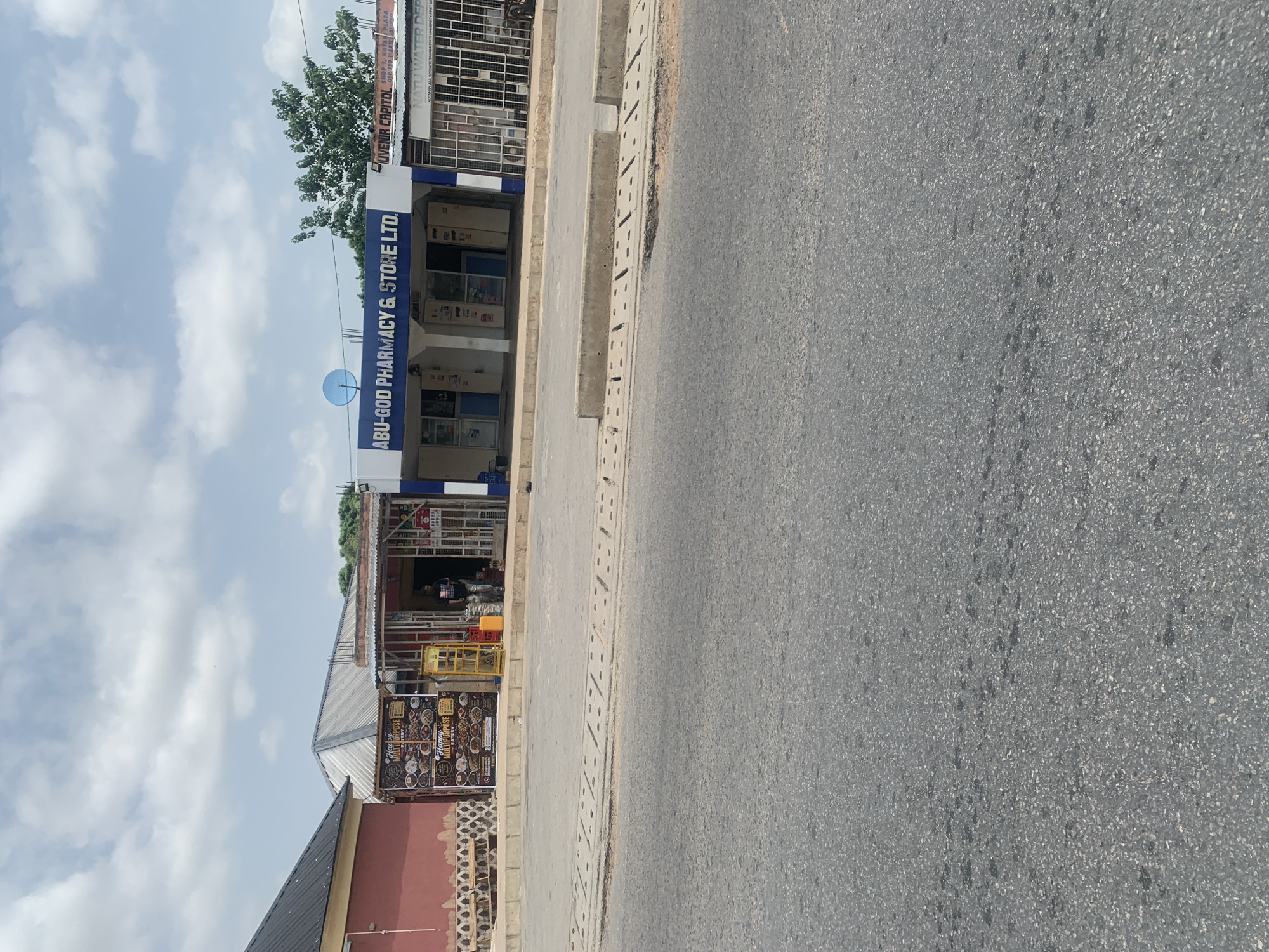 The image shows a street scene in Jalingo, Nigeria, under a partly cloudy sky, suggesting it is daytime. The perspective is from a low angle, looking up and to the right along a paved road. On the left side of the road, a row of shops with blue and white awnings lines the street. One prominent shop has the text "ABU-GOD PHARMACY & STORE LTD." in large blue letters. Other visible text on signs above the shops includes "GUBERN CAPITOL", "KETU WATER DEP.", and "Happy Multipurpose Eatery". A satellite dish is visible on the roof of the pharmacy.
To the right of the shops, a concrete sidewalk with drainage channels runs parallel to the road. The road itself is asphalt, with some dark patches and marks, possibly from oil or water. The scene appears to be a commercial area, with shops indicating a bustling local economy. There are no distinct signs of a specific event or activity taking place, and the overall impression is a typical day in a Nigerian town. The image is oriented vertically, with the sky at the bottom and the ground at the top, suggesting it was taken with the camera rotated.